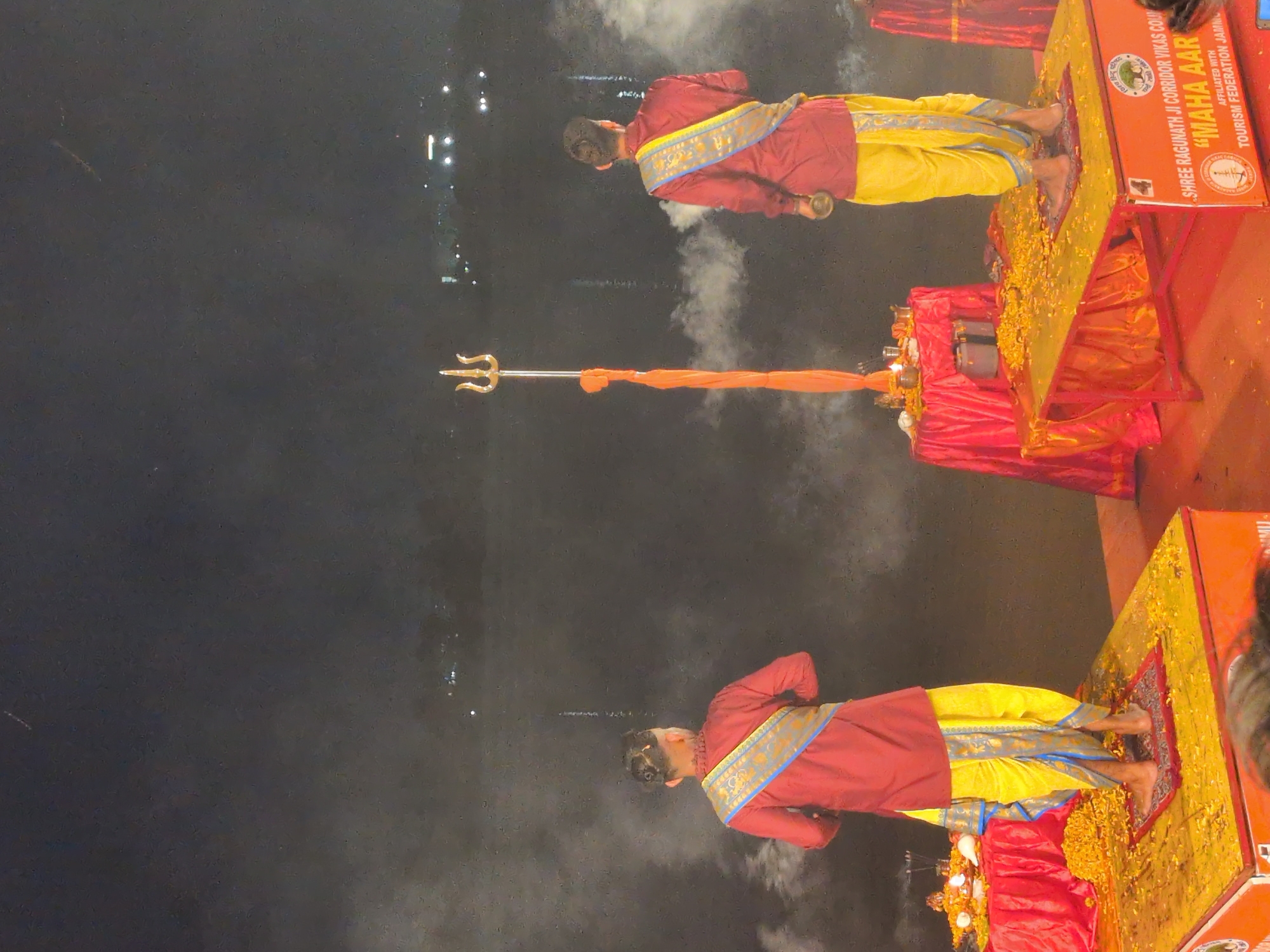 The image depicts a religious ceremony taking place at night, likely on the banks of the Tawi River in Jammu, India, given the presence of the "Shree Raghunath Ji Corridor Vikas Council" and "Maha Aarti" text visible on a structure. Two men, dressed in traditional Indian attire consisting of maroon kurtas and yellow dhotis with decorative blue and gold sashes, stand on raised platforms. Both men appear to be participating in a ritualistic offering or prayer, with one holding a small metal vessel. In the center of the scene, a tall trident, or 'trishul', is erected, adorned with a vibrant orange cloth. Smoke or mist is present around the figures and the river in the background, creating a slightly ethereal atmosphere. The platforms are decorated with yellow flowers, and red fabric drapes are visible. The scene is illuminated by artificial lights, highlighting the ceremonial aspects.