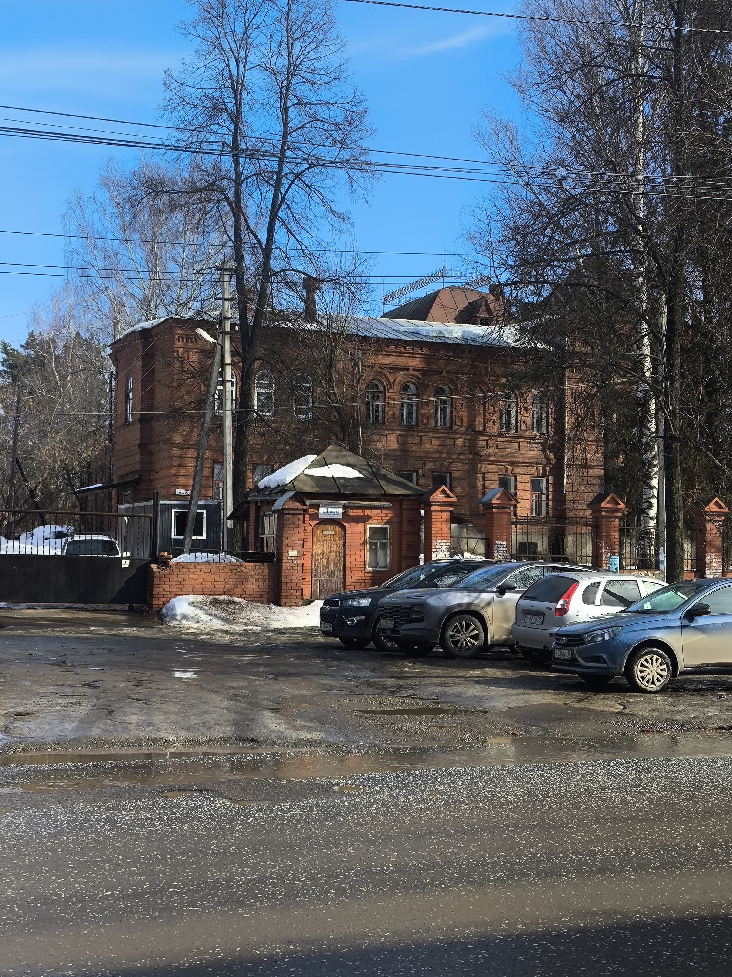 The image depicts a street scene in Votkinsk, Russia, featuring a two-story brick building with architectural details, a small gatehouse, and parked cars. The building is red-brick with arched windows on the upper floor and a dark metal roof. A utility pole with wires is visible in front of the building. Several cars are parked in front of the building, including a blue sedan, a silver hatchback, a gray SUV, and a dark SUV. The road is wet with puddles and patches of ice or snow. Bare trees surround the scene, and the sky is bright blue with a few wispy clouds.