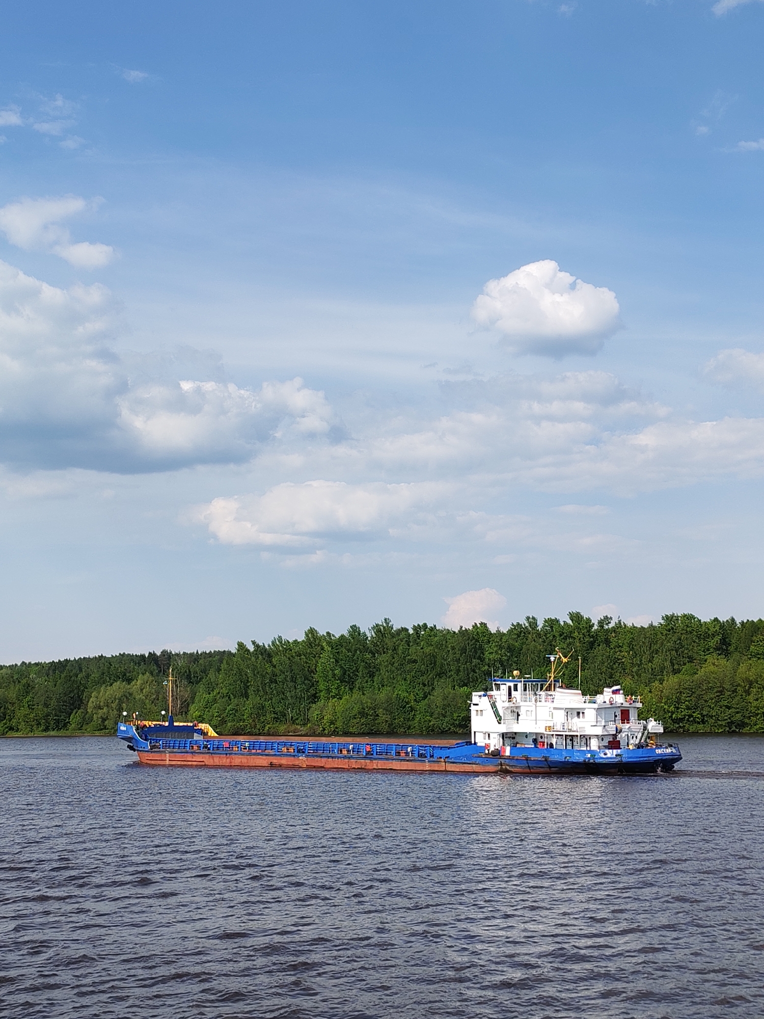 A barge navigates a body of water. The barge is painted in shades of blue, brown, and white. Lush green trees line the riverbank in the background. The sky overhead is blue, dotted with fluffy white clouds. This image was captured in Ermolino, Russia.