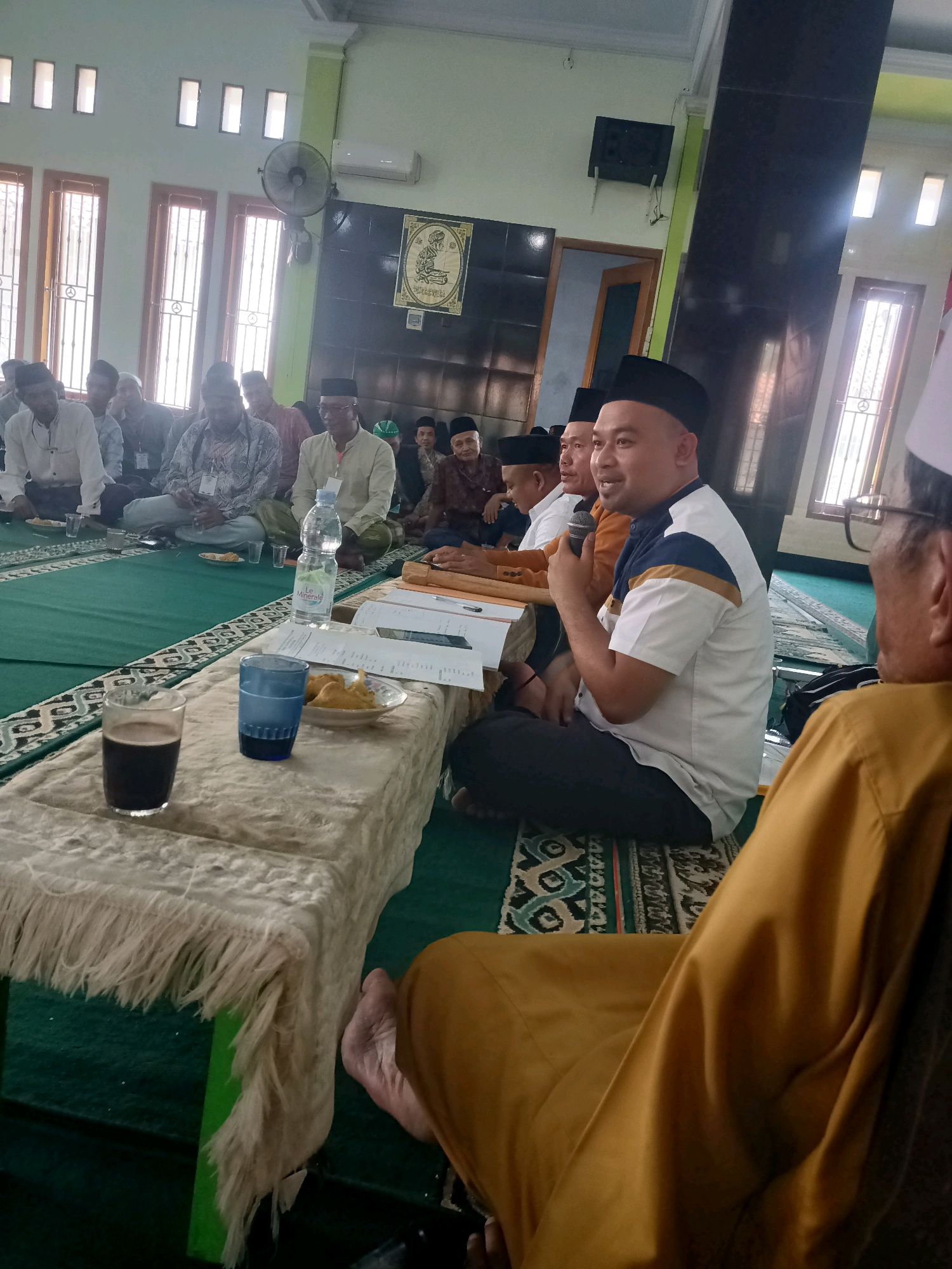 The image depicts an indoor gathering, likely in a mosque in Penusupan, Indonesia. Several men are seated on a green carpet, some wearing traditional Islamic attire, including peci hats. In the foreground, a table is draped with a fringed cloth, holding a glass of dark liquid, a blue glass, a plate of fried snacks, and some papers. One man in the mid-ground, wearing a white shirt with blue and gold accents and a black peci, is speaking into a microphone. Other individuals appear to be listening or engaged in conversation. The room has tall windows with vertical slatted blinds and fluorescent lighting on the ceiling. A ventilation fan is visible on the wall. The overall atmosphere suggests a religious or community meeting.