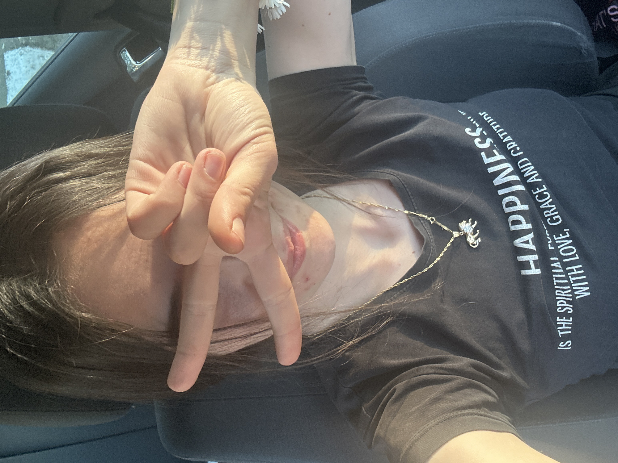 A woman is in a vehicle, presumably a car, obscuring part of her face with her right hand, with fingers outstretched in a "V" formation. She is wearing a dark t-shirt with a white inscription stating, "Happiness is the spiritual fuel with love, grace and gratitude". She is also wearing a gold necklace with a pendant. A white flower appears to be pinned to her left arm. The interior of the vehicle is visible, including the car door and seat. The location cannot be determined to be Resen, Bulgaria with the information provided in the image.