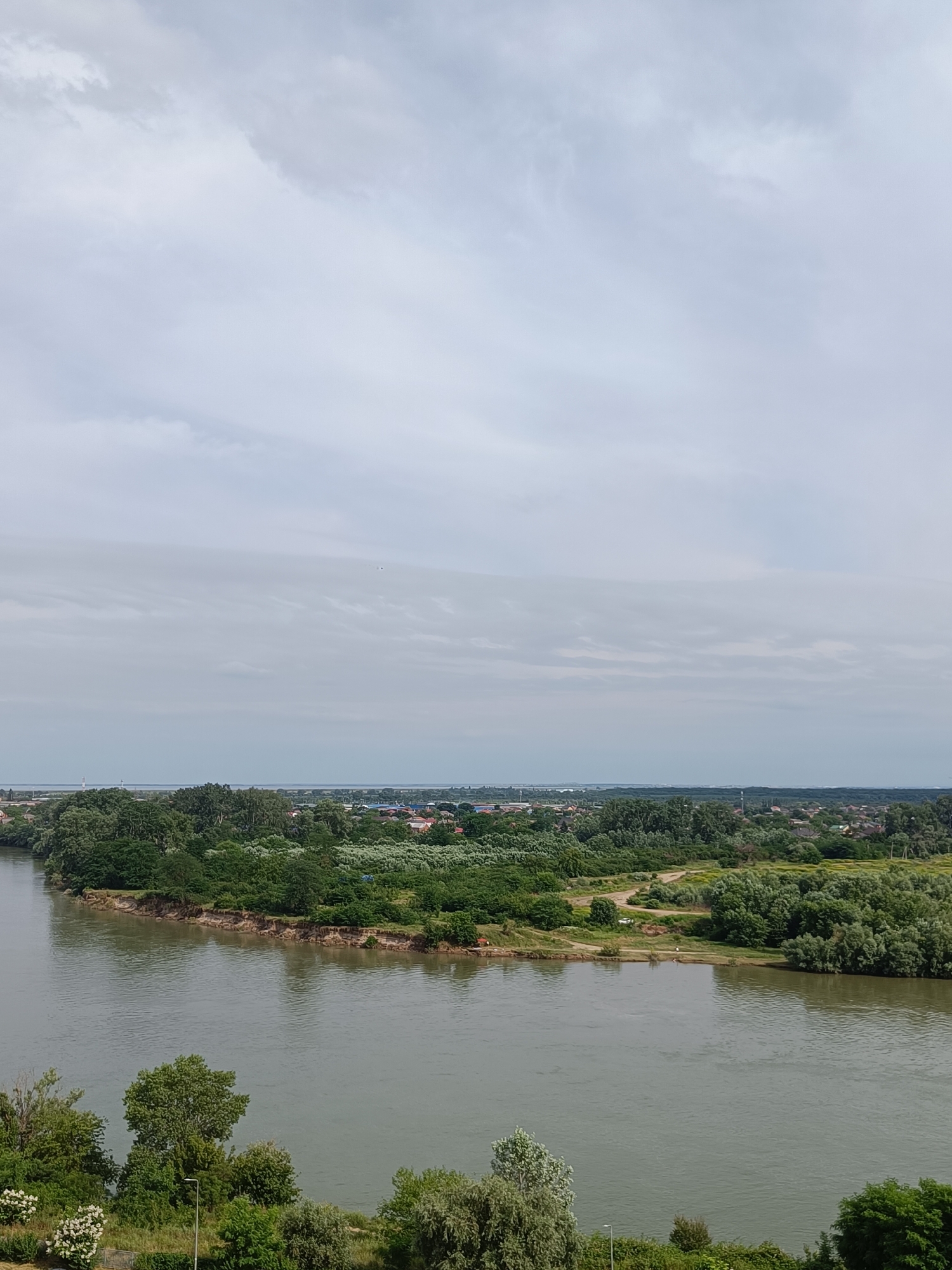 An aerial, wide shot captures a wide, calm river in the foreground, reflecting the overcast sky. The river curves from the lower left to the middle right of the frame. The left bank of the river is lined with lush green trees and vegetation. The right bank features a peninsula of land covered in dense green trees and shrubbery, with a small clearing and a dirt path visible. In the distance, beyond the river and vegetation, a sprawling town or city is visible, with rooftops and structures peeking through the trees. The sky is filled with diffused grey clouds, indicating an overcast day. The image appears to be taken from a high vantage point, possibly a hill or tall building. The overall scene depicts a natural landscape meeting a populated area. Based on the distinctive bend of the river and the presence of a large, nearby city, this view is likely of the Kuban River at its confluence with the Karasun River, near the city of Krasnodar, Russia.