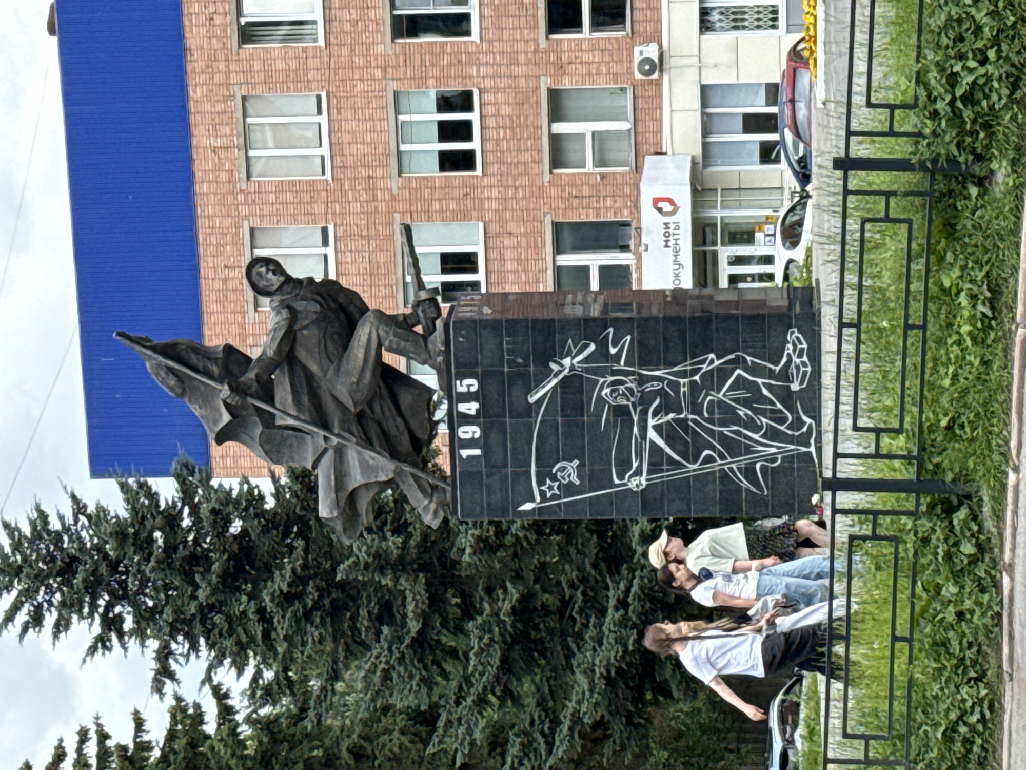 In Izhevsk, Russia, a war memorial stands prominently in front of a brick building topped with a blue roof. The memorial itself is a dark, monolithic structure featuring a dynamic bronze sculpture of a soldier raising a flag, with the year "1945" inscribed on its face. Below the sculpture, a white outline drawing depicts another soldier with a flag and a Soviet star. Three young women, dressed casually in t-shirts and pants, walk past the memorial. The environment includes lush green grass and trees, with a black metal fence separating the sidewalk from the foreground. In the background, parked cars and a sign that reads "МОИ ДОКУМЕНТЫ" (My Documents) are visible.