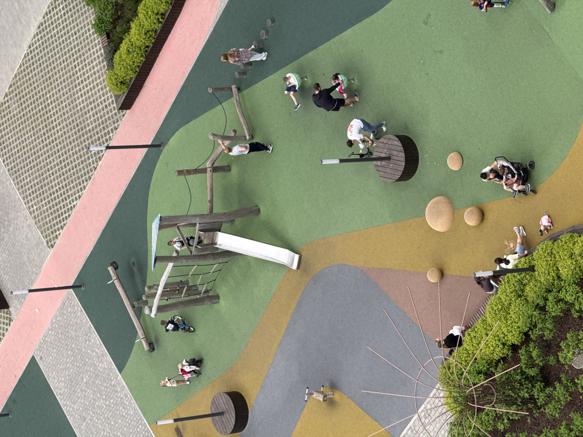 The image shows an aerial view of a playground. The playground is situated within a city, likely Perm, Russia, given the context. The ground is covered in various colored synthetic materials, including green, yellow, gray, and brown. The playground equipment includes a slide, climbing structures made of logs and ropes, and wooden platforms. Several people are present on the playground, including children playing on the equipment and adults accompanying them. Some people are sitting on benches near greenery. The surrounding area features paved surfaces in a grid pattern and patches of green vegetation. Several light posts are distributed throughout the playground.
