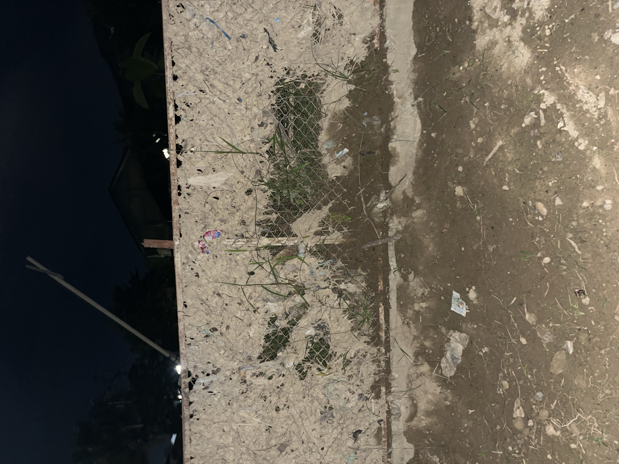 The image depicts a section of a dilapidated fence at night in Kota Kuala Simpang, Indonesia. The fence appears to be constructed from mesh wire covered with a layer of decomposing organic material and what seems to be shredded paper or fabric. Various pieces of debris, including plastic wrappers and small pieces of trash, are embedded within and hanging from the fence. Green grass and weeds are growing through and around the fence. The ground in front of the fence is muddy and littered with more debris, including plastic bags and small stones. In the background, silhouetted trees and buildings are visible against a dark night sky. A wooden pole extends diagonally upwards from the left side of the frame, intersecting with the sky. The scene suggests an area that is neglected and possibly a dumping ground.
