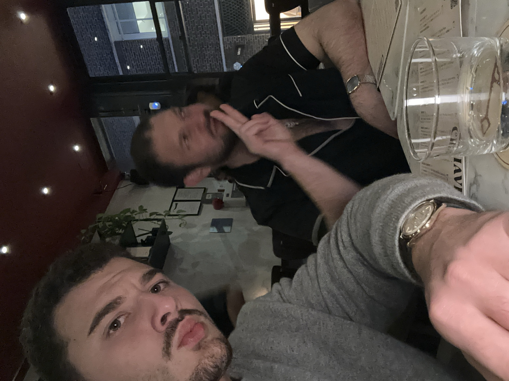 The image depicts an indoor scene at night within a modern restaurant or bar located on an urban street in London, United Kingdom.
In the foreground, an adult male with dark hair, a mustache, and a short beard wears a grey long-sleeved sweater and a gold-toned wristwatch on his left wrist. He is positioned for a selfie, looking directly at the camera with a neutral expression.
Seated behind and to the right of the foreground subject is a second adult male with dark hair, styled into a ponytail or bun, and a beard and mustache. He is wearing a black short-sleeved collared shirt with white piping, unbuttoned to reveal his chest and a silver-toned necklace. A silver-toned wristwatch with a mesh strap is visible on his left wrist. His right hand is raised, forming a "peace sign," and he is looking off to his right, away from the camera.
The two men are seated at a table with a white marble-patterned surface. A clear glass filled with water rests on a coaster in the lower right foreground, positioned over a paper placemat or menu with partially visible text, possibly "CHAVI" or "MAVI" in reverse. Additional paper items are visible on the table.
The interior environment features a dark reddish-brown ceiling with multiple recessed spotlights emitting warm light. The walls are light grey and appear to have a textured finish. Dark shelves adorned with small potted plants are visible on the left wall.
Through a large window or glass door in the background, an exterior urban scene is visible, illuminated at night. A dark brick building with rectangular windows and white frames is prominent. A partial sign is visible on the exterior building, appearing to read "SOUT" or "SOUL."