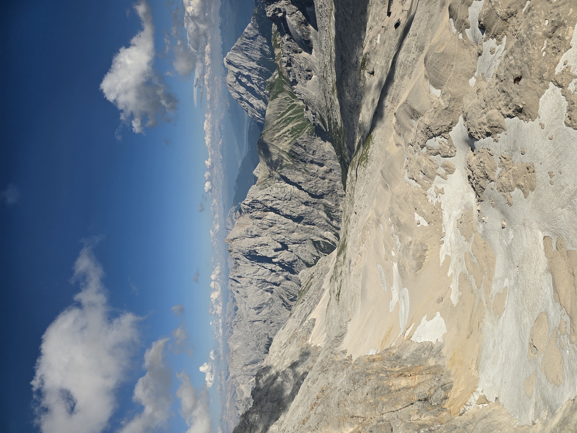 The image is a high-angle shot of a vast mountain landscape, likely taken from a high vantage point. The foreground is dominated by rocky, sandy terrain with patches of snow or ice. The terrain slopes downwards and towards the right. In the middle ground, rugged, grey mountains rise steeply, with some showing patches of green vegetation on their slopes. Further in the background, a range of more distant, softer-looking mountains fade into the haze under a bright blue sky with scattered white clouds. The scene appears to be a natural, mountainous environment, potentially a ski resort area or a hiking destination. There are no discernible people or specific man-made structures visible in detail, although there may be very small structures or ski lifts in the far distance. The overall impression is one of a grand, natural, and expansive alpine vista. Given the context of Garmisch-Partenkirchen, Germany, this scene is highly likely to be the Zugspitze or a similar high mountain area.