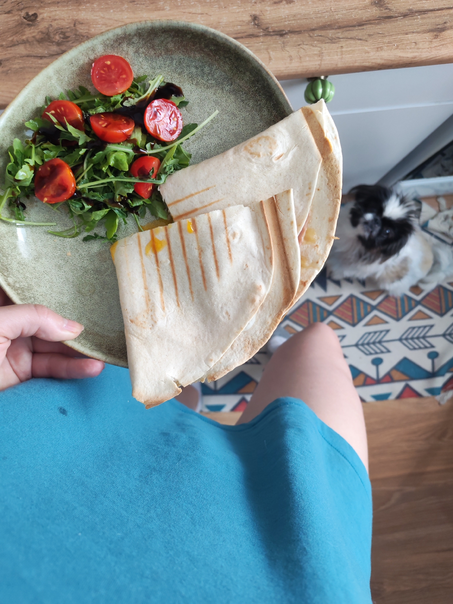 A close-up, top-down perspective shows a person's hand holding a plate of food. The plate contains a salad of arugula and halved cherry tomatoes drizzled with a dark sauce, and several folded quesadillas with grill marks. To the right, out of focus, a small dog with black and white fur sits on a patterned rug. The background includes a wooden surface, a white cabinet with a green knob, and the floor. The image appears to be taken indoors, likely in a kitchen or dining area. No specific location in Izhevsk, Russia can be determined from the visual information provided.