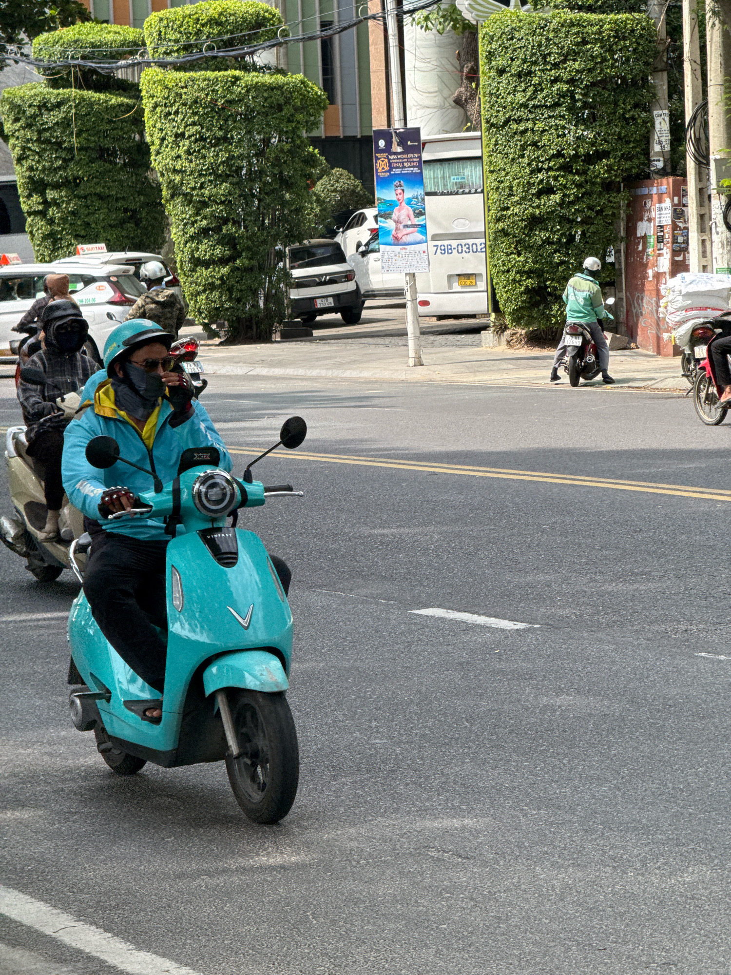 A person rides a turquoise electric scooter on a paved road in Nha Trang, Vietnam. The rider wears a blue jacket, yellow shirt, dark pants, a teal helmet, a black face mask, and gloves. The scooter has a white "VINFAST" logo on the front. Another person on a light-colored scooter is visible behind the first rider, wearing a plaid jacket and a black hood. The street is lined with tall, manicured green hedges. In the background, a white bus with "79B-030.26" on its license plate is parked near a pole with advertisements. Several cars and motorcycles are also present on the road and sidewalk.