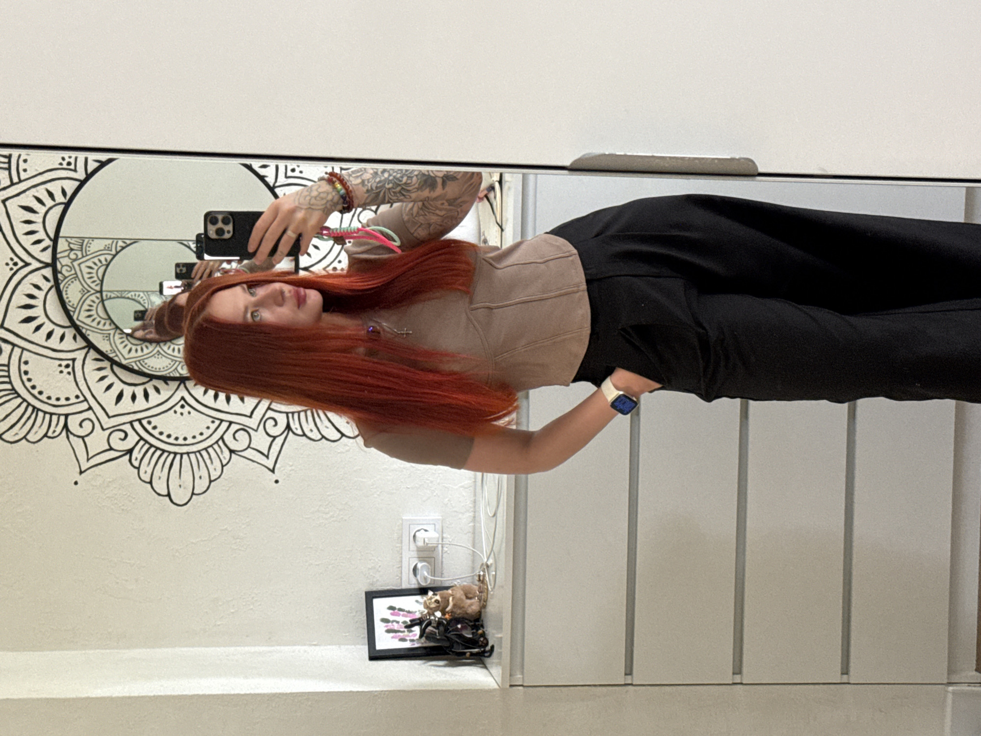 A young woman with long, straight, red hair is taking a selfie in a mirror. She is wearing a tan-colored top and black pants. The mirror reflects the woman's face, upper body, and the hand holding the phone. Behind her, a mandala wall decoration is visible. Below the mandala, an outlet and a framed picture on a shelf can be seen. The location is likely inside an apartment or house; however, there is no evidence to confirm the city of Rostov-on-Don, Russia.
