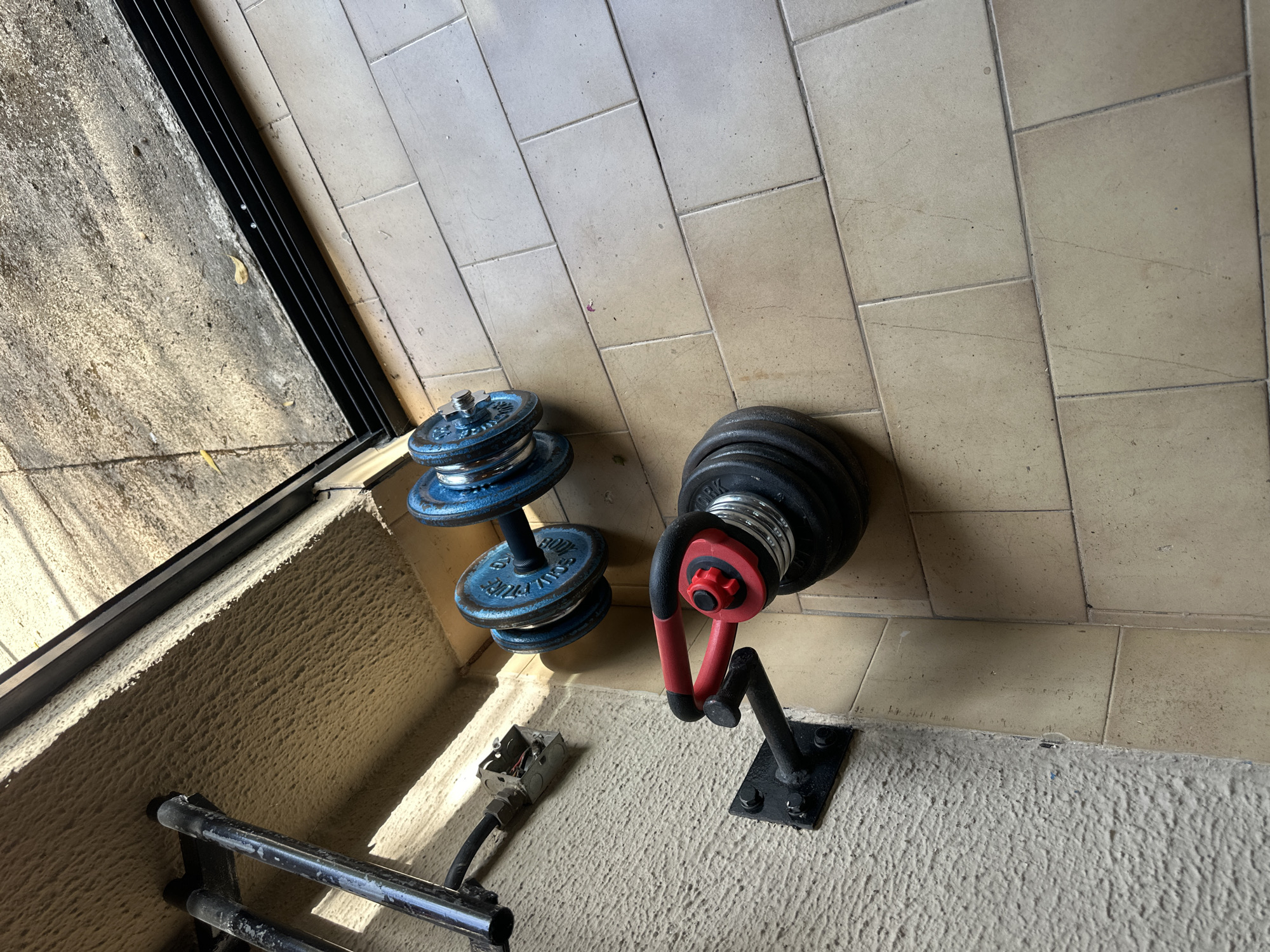 The image shows two sets of dumbbells, one blue and one black, resting on the floor of an indoor/outdoor space. The blue dumbbell sits directly on the floor, while the black dumbbell is held in a black and red wall-mounted dumbbell rack. The floor is tiled with beige square tiles. The adjacent wall is textured and painted white. There is an electrical box with exposed wires and a section of a metal railing visible along the bottom of the image. The location of the image in Jos, Nigeria is not known.