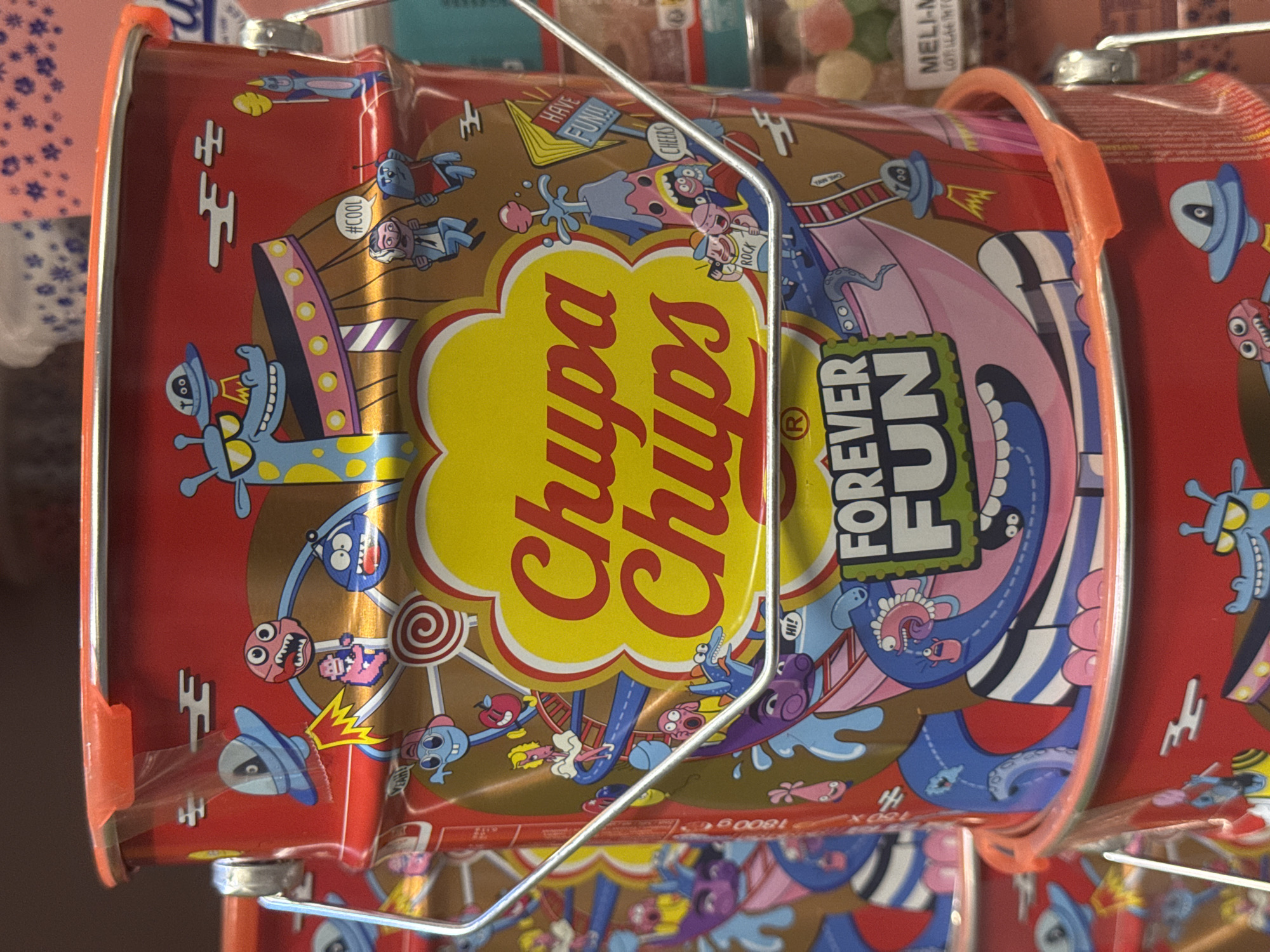 The image shows a close-up of a red Chupa Chups tin filled with candies. The tin is decorated with a colorful, cartoonish design featuring various characters, including aliens, monsters, and abstract shapes. The Chupa Chups logo is prominently displayed in yellow and red on the front of the tin. The tin also has an orange plastic handle and a metal wire frame attached. In the background, other containers of candy are visible, suggesting a retail or storage setting. The overall impression is vibrant and playful, reflecting the brand's association with fun and sweets. The text on the tin includes "Chupa Chups," "FOREVER FUN," "#COOL," "HAVE FUN!!," "ROCK," and "CHEERS." Some of the background text is also visible, including "1800 ge" and "180X." The location context is Vilvoorde, Belgium, though this is not explicitly visible in the image itself.
