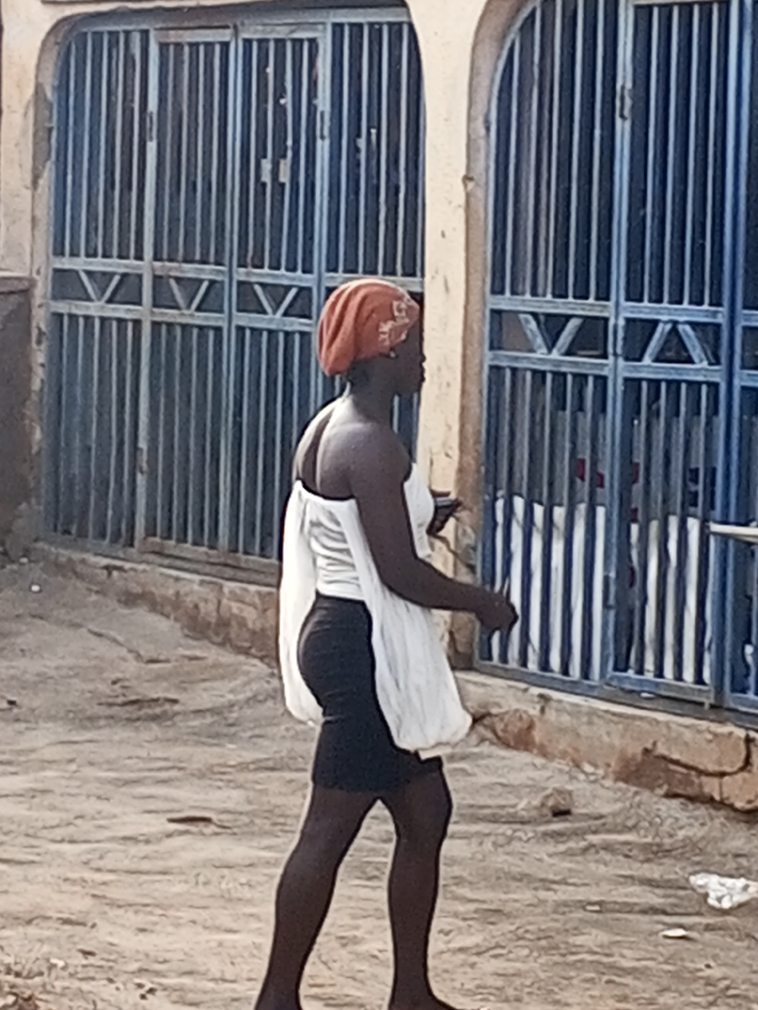 A person walks barefoot on a dirt road in Gwong, Nigeria. They are wearing a red head covering, a white top, and a black skirt or shorts. Behind the person is a building with blue metal doors with a "V" design on the bottom. The building appears to be made of stone. The ground is a light brown color.