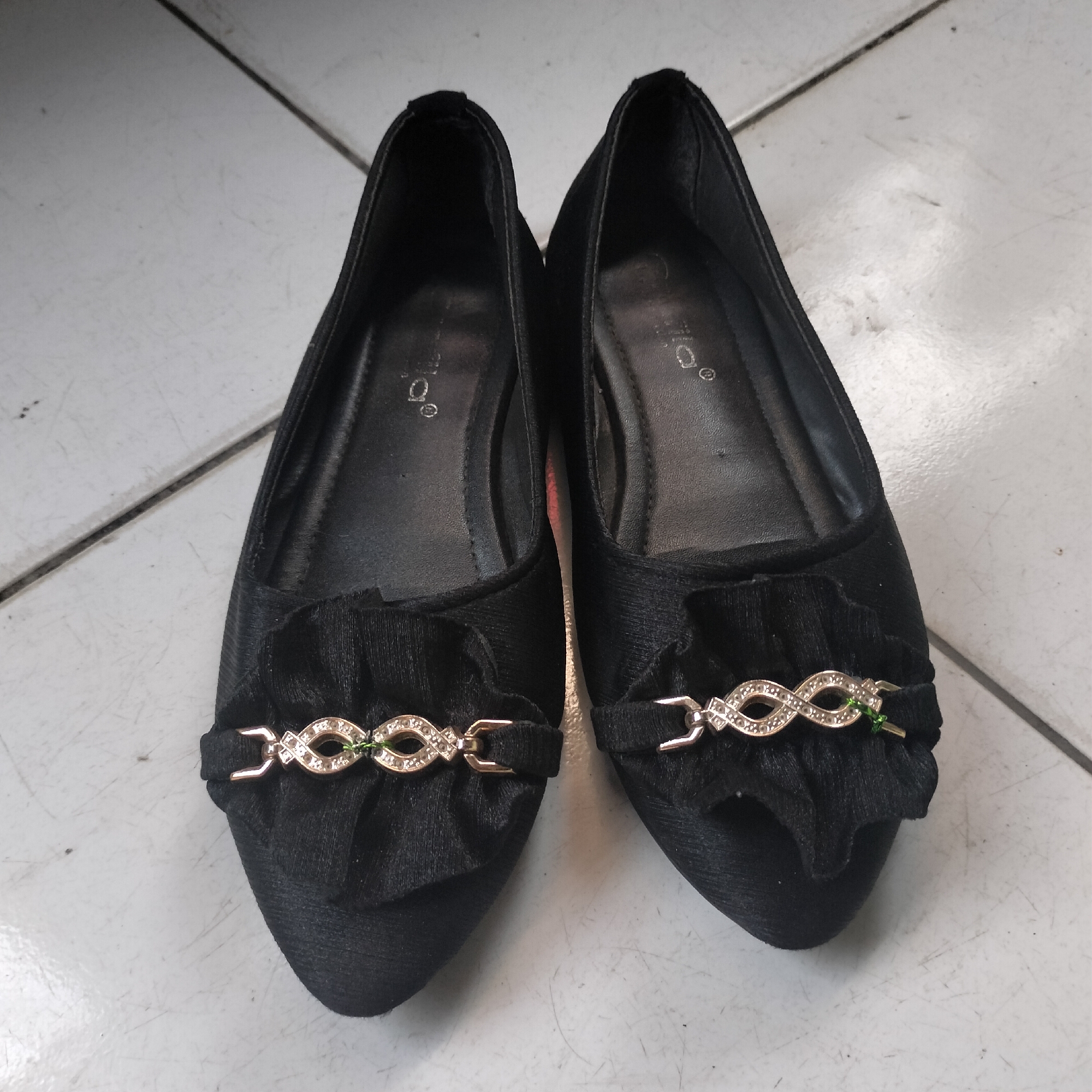 A pair of black flat shoes with a ruffled detail and ornate buckle embellishments are placed on a tiled floor. The buckle on each shoe features a gold-colored infinity symbol adorned with clear rhinestones and a small green jewel. The shoes appear to be made of a textured fabric, possibly velvet or a similar material. The tiled floor is light gray with darker grout lines forming a grid pattern. The lighting suggests an indoor environment, possibly a home. The image was taken in Tangerang, Indonesia.