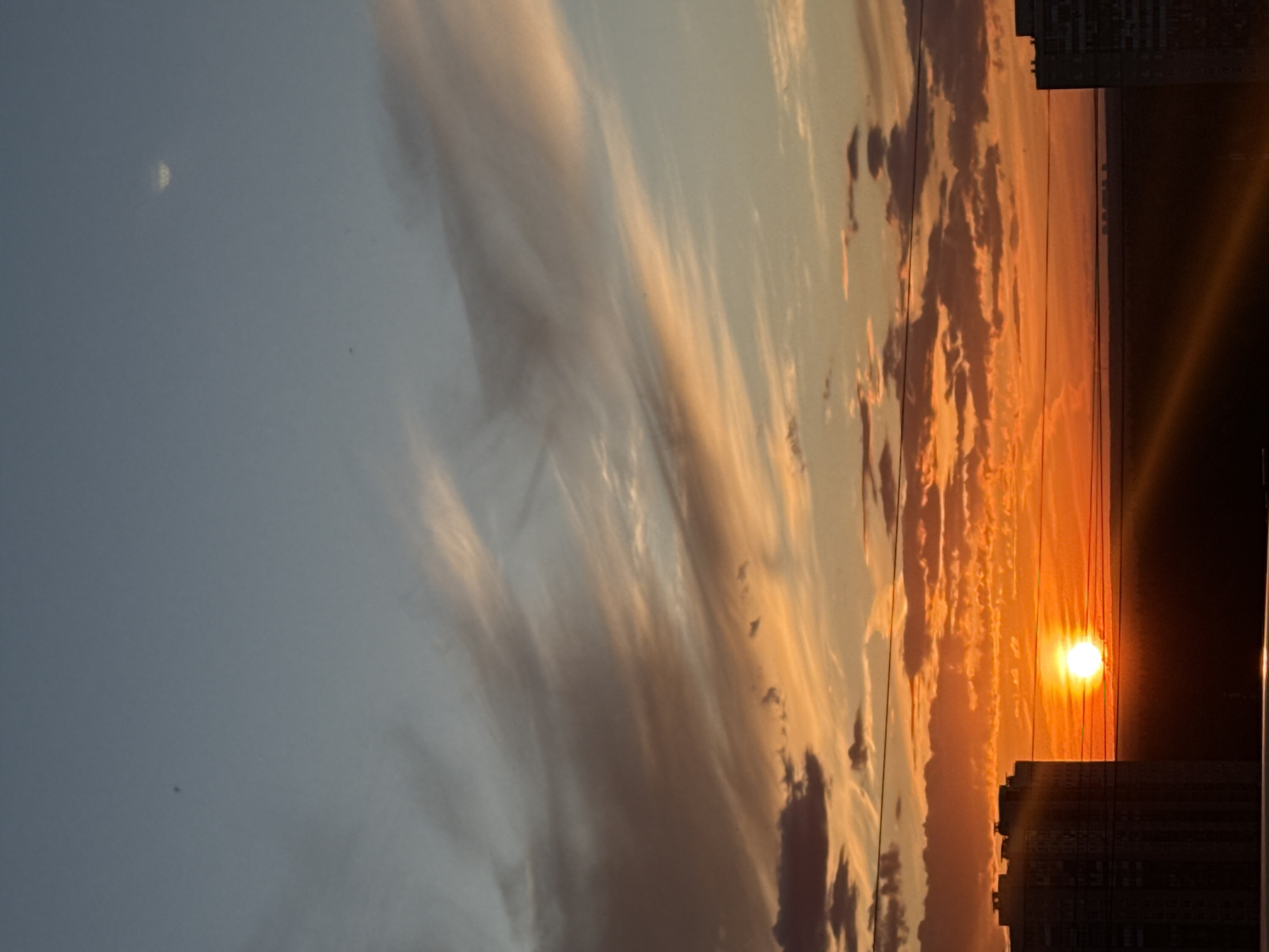 The image depicts a sunset over the city of Khimki, Russia, with a view of apartment buildings and the sky filled with clouds. The sun is low on the horizon, casting a warm orange glow across the clouds. The sky transitions from a pale blue at the top to vibrant orange and red hues near the horizon. Two tall apartment buildings are visible on the left and right sides of the frame, silhouetted against the colorful sky. The building on the left is closer and more detailed, showing the structure of windows and balconies. The building on the right is further away and less distinct. The landscape between the buildings is dark and appears to be a natural area or perhaps more distant buildings. The scene is captured from a high vantage point, suggesting an elevated view of the city. The overall atmosphere is serene and peaceful, characteristic of a sunset.
