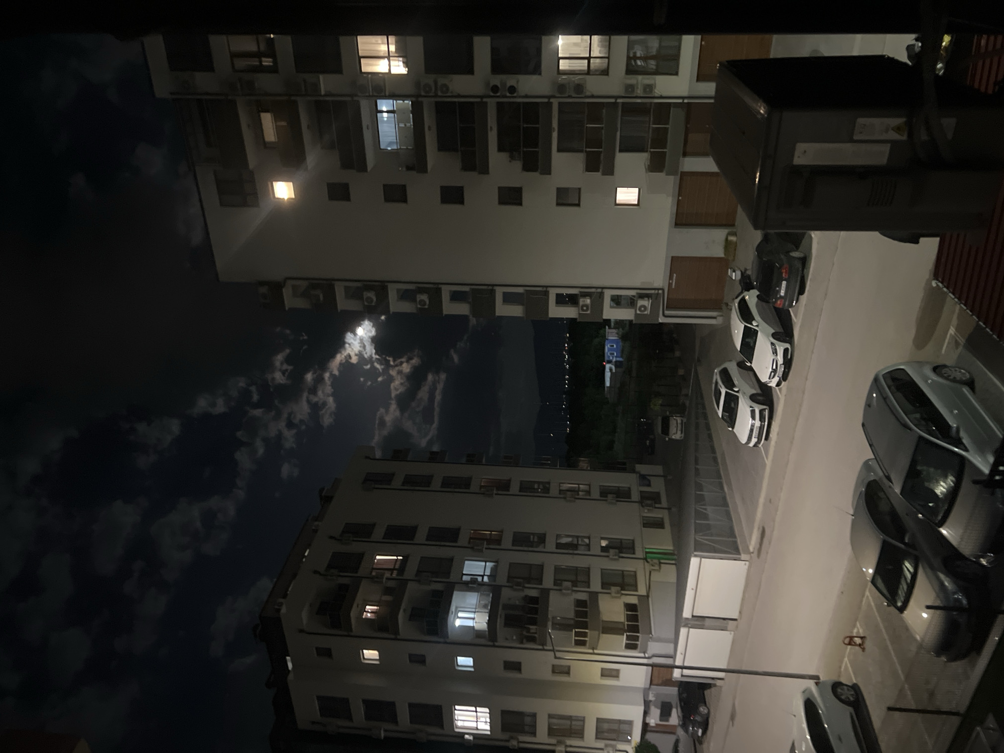 At night, a view from an elevated position shows two white apartment buildings in Plovdiv, Bulgaria. The buildings have multiple floors with rows of windows and air conditioning units. A parking lot between the buildings contains several parked cars. The night sky is visible, with the moon partially obscured by clouds. A street with lights is visible in the distance between the buildings.