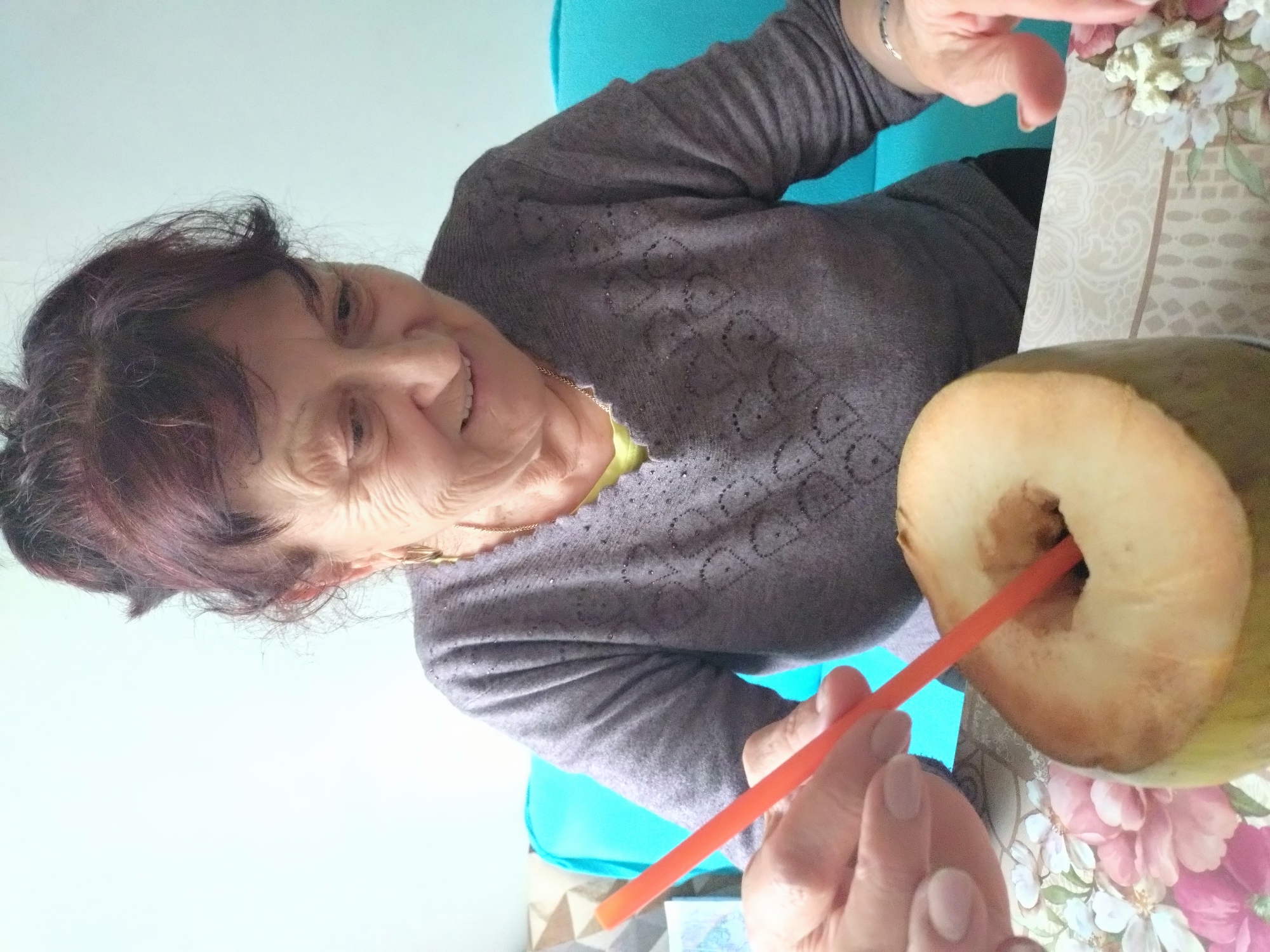 A middle-aged woman with short, dark hair, wearing a long-sleeved gray shirt with a decorative pattern, is seated at a table. She is smiling and holding an orange straw. A coconut with a hole cut in the top, through which the straw is inserted, is also on the table. The table has a floral patterned tablecloth. She is sitting on a blue cushion. The location is likely indoors in Stara Zagora, Bulgaria, though there's no specific indication to verify this.