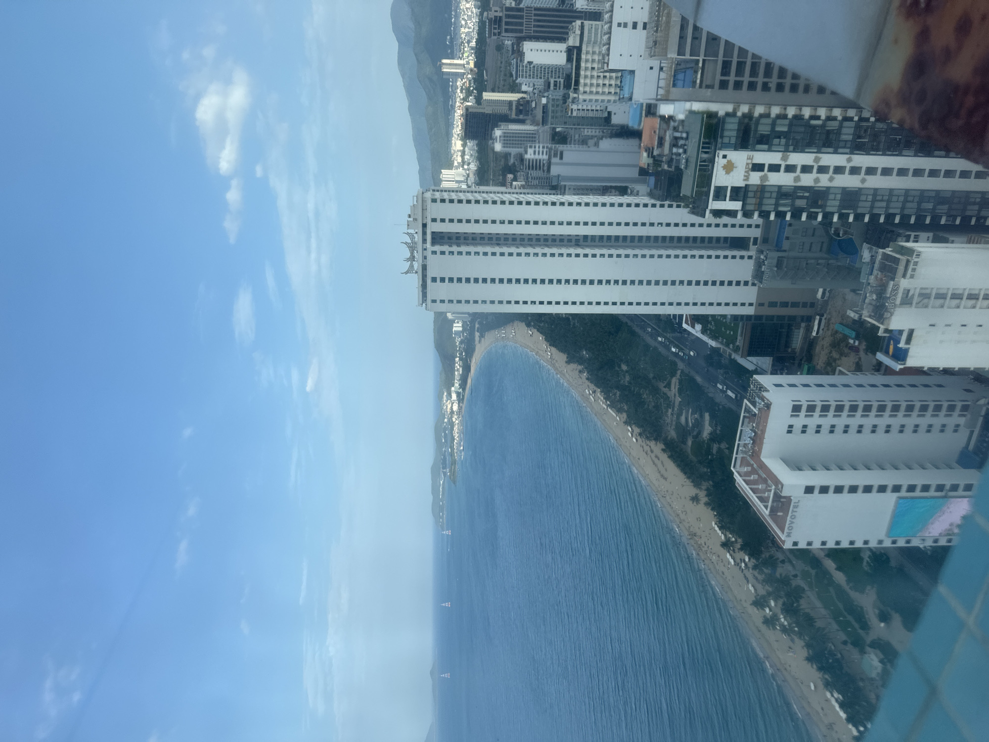 The image presents a high-angle, panoramic view of a vibrant coastal city, likely taken from a tall building or observation deck. The sky is a clear, bright blue with scattered white clouds, indicating fair weather during the daytime.
The prominent features of the scene are a long, curved sandy beach and the expansive blue ocean. The ocean waters appear calm with gentle ripples, and several small boats or buoys are visible at various distances from the shore. The beach itself is lined with palm trees and appears to have numerous sun loungers or beach umbrellas. A road runs parallel to the coastline, separating the beach from a dense urban landscape.
The city is characterized by numerous high-rise buildings, many of which are modern and multi-storied. One large white building along the beachfront prominently displays the text "NOVOTEL" and features a visible swimming pool. Other buildings further inland bear names such as "MAPLE" and "BOSS." These structures extend significantly inland, reaching the base of a range of mountains that form the backdrop in the distance. The overall impression is that of a bustling, developed coastal resort city. A blurred blue tiled surface in the bottom right corner suggests the vantage point was from a building with a balcony or window.