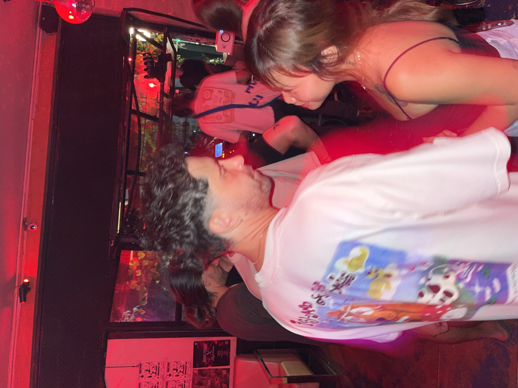 The image shows an indoor scene, potentially a bar, with red lighting. A man with curly hair is wearing a white t-shirt with a graphic design on the back. A woman with dark hair and a spaghetti strap top leans toward him. Other people are in the background. A disco ball hangs from the ceiling. A window shows greenery outside. Posters are visible on a wall. The location is North Kuta, Indonesia.