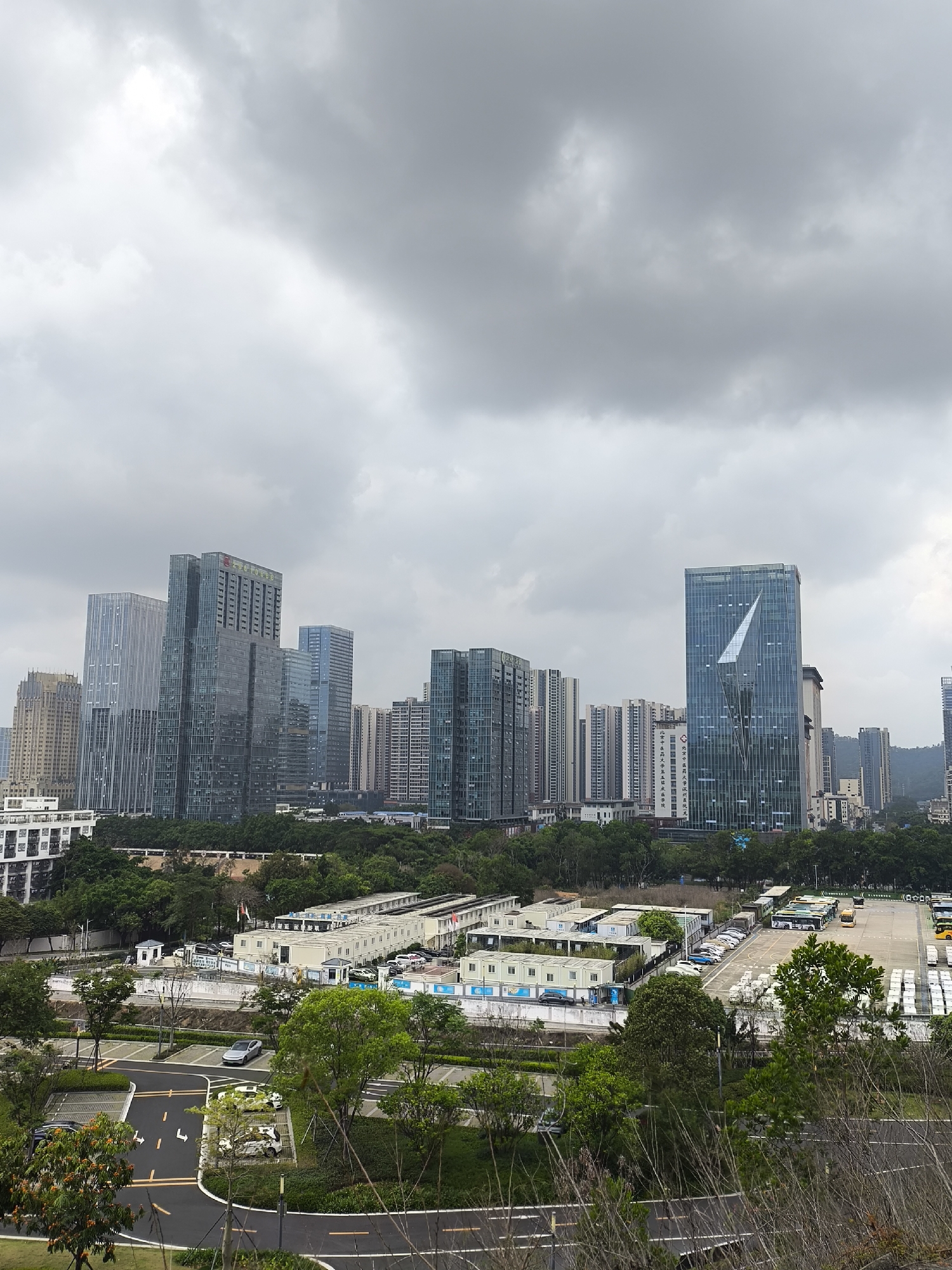 The image shows a view of the Longgang District, Shenzhen, China. In the foreground, there is a road with yellow lines, followed by greenery consisting of trees and grass. Behind these, rows of low, rectangular buildings are visible, possibly temporary structures or modular homes. Further back, a line of trees separates the low buildings from the cityscape. The skyline is dominated by a collection of modern high-rise buildings with glass facades, some taller than others. The sky above is overcast with heavy gray clouds. There are no visible people in the image. The scene appears to be taken from a higher vantage point, overlooking the area.