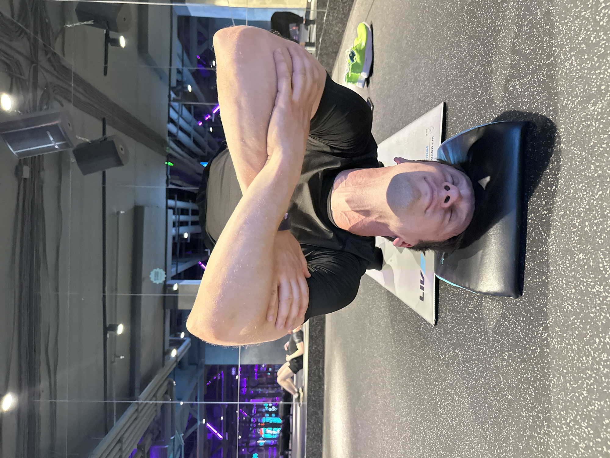 This media file shows a man lying on his back on a mat, appearing to be in a gym or fitness studio. He is wearing a black t-shirt and black shorts. His arms are crossed over his chest, and his head is resting on a black mat. The setting features a speckled gray floor, characteristic of gym flooring. In the background, a large reflective surface, likely a mirror or glass wall, shows a reflection of the gym's interior, including lighting fixtures, cables, and what appear to be other people engaged in exercise, some illuminated by purple lights. A bright green sneaker is visible on the floor to the right of the man. The overall impression is that of a workout session in progress. There is some text visible on the mat that appears to read "LIV" and potentially other lettering that is partially obscured. The lighting suggests it could be daytime, though indoor lighting is predominant.