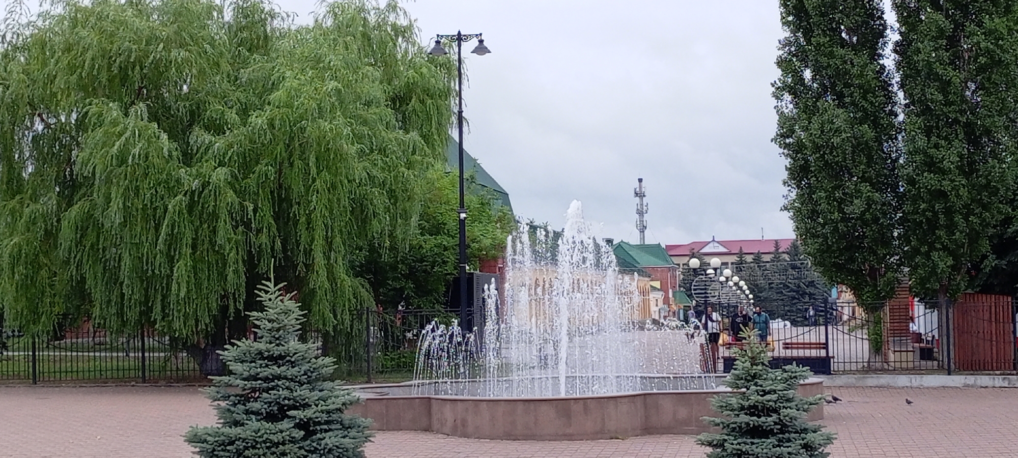 The image shows a public park or square in Kirsanov, Russia, on an overcast day. A large weeping willow tree dominates the left side of the frame, with its long branches reaching towards the ground. In the center, a multi-tiered fountain sprays water upwards. To the right of the fountain, two young evergreen trees are visible. Behind the fountain, several people can be seen, some standing and others walking, near buildings with red and green roofs. A tall, slender street lamp with two lights stands to the left of the fountain, and another similar lamp is partially visible on the far right. The ground is paved with brickwork. In the background, a cellular tower and various trees, including tall, dark green trees on the right, complete the scene.