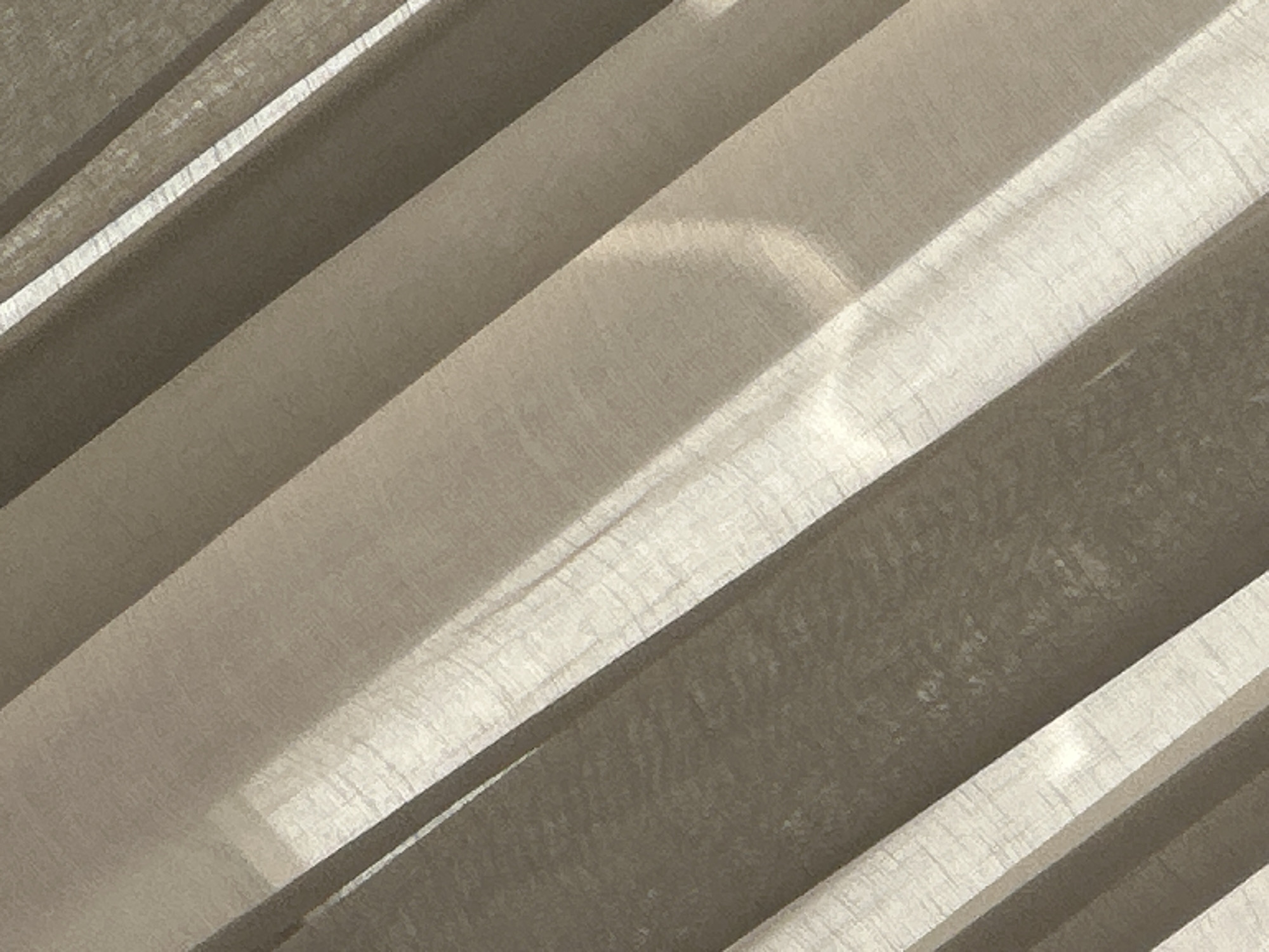 The image displays a close-up, abstract view of sheer fabric curtains. The curtains are oriented diagonally, creating alternating bands of light and shadow. The fabric appears textured, with a visible weave. The lighting suggests the presence of a light source, possibly sunlight, filtering through the material, casting soft shadows and highlights. There are no people, animals, or discernible objects in the scene. The background is not visible. The image provides no information that would allow for its localization to Mañğıstaw, Kazakhstan, or any specific urban environment.