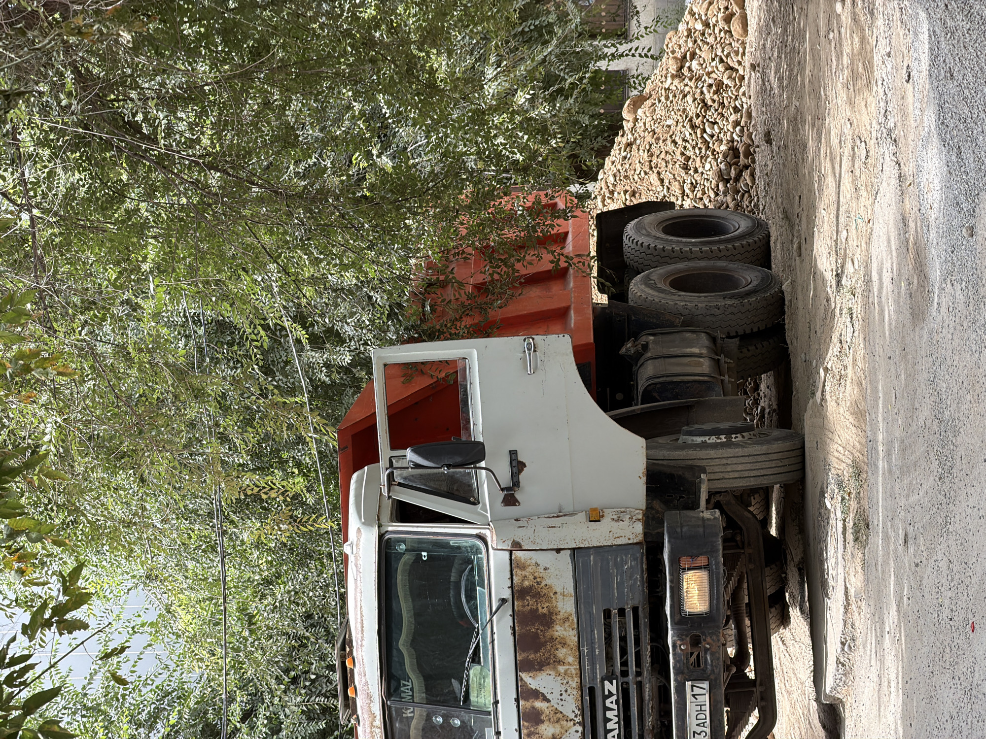 An aged, white and orange dump truck, possibly a KAMAZ model given the visible badging, is parked on a dirt road in Shymkent, Kazakhstan. The truck exhibits significant rust and wear, particularly on its front white panels and the driver's side door, which is ajar. The truck bed is bright orange and appears empty, situated next to a large pile of rounded, light brown stones and gravel. Lush green foliage from trees overhangs the scene, casting dappled shadows on the truck and the ground. The ground surface is a mix of packed dirt, loose gravel, and sand. The truck's headlights and license plate are visible, the plate displaying characters that are partially obscured. The overall scene suggests a construction or landscaping site. There are no people visible in the image.