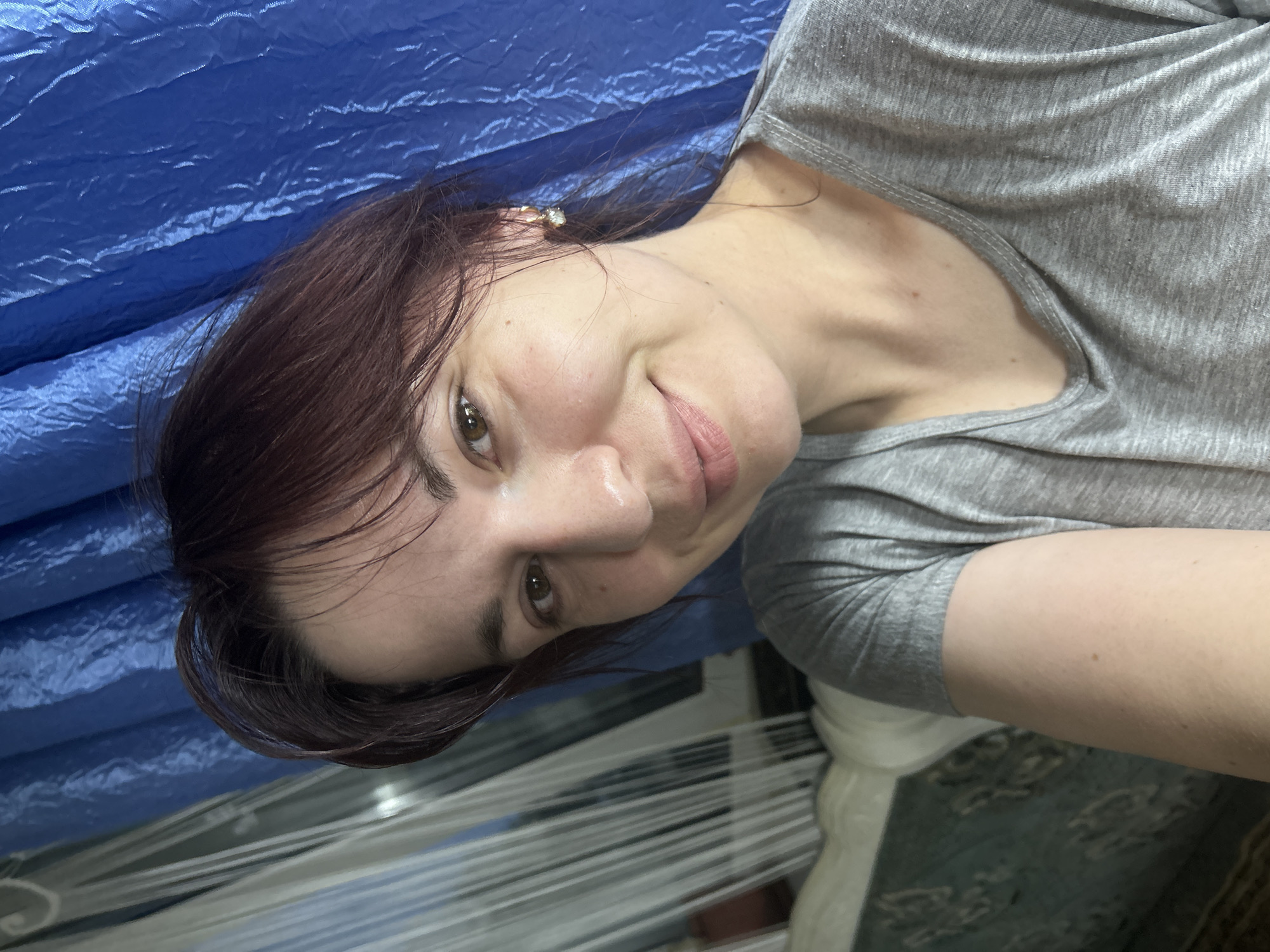 A selfie taken indoors shows a woman with shoulder-length, dark brown hair, styled loosely with some strands falling across her forehead. She is wearing a grey t-shirt and a small earring with a crystal in her right ear. Her skin is fair with some natural blush on her cheeks. She has brown eyes and is smiling gently. The background behind her is a blue, crinkled fabric, possibly a curtain or sheet, and to her left, a white, sheer fabric with vertical lines is visible, which might be another curtain or a mosquito net. Part of a light-colored sofa with a patterned textile is also visible in the lower left of the frame. The image appears to be taken in a residential setting, likely a bedroom or living room. There is no identifiable information to suggest a specific location in the city of Fokino, Russia.
