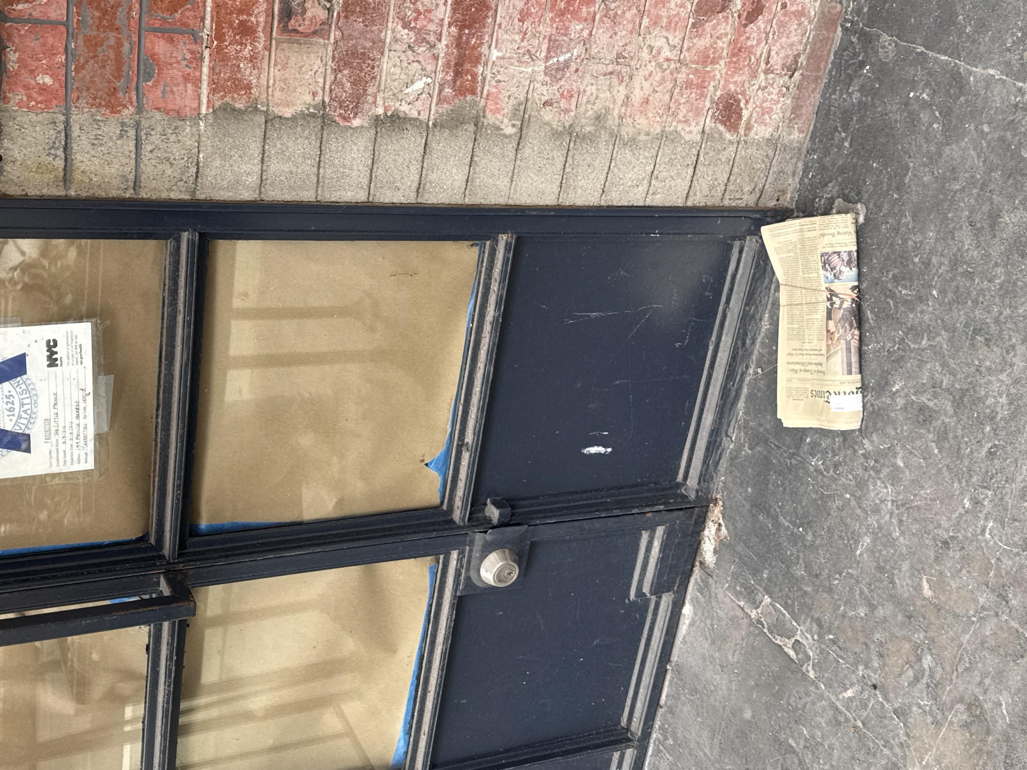 The image shows a doorway in New York City. A blue-framed door with glass panels is set against a brick and stone wall. A newspaper, identified as the "New York Times", lies on the ground next to the door. A partially obscured NYC Health inspection card is taped on the left-most window. The inspection card identifies the location as "THE LITTLE PRINCE", 199 PRINCE STREET, in Manhattan.