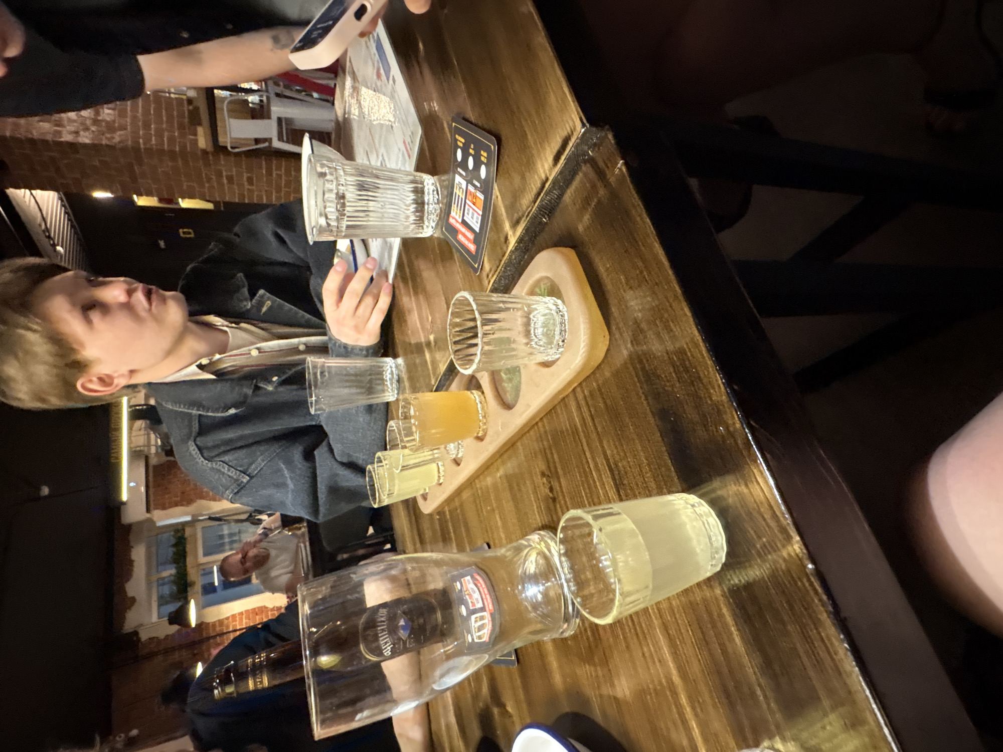 A dimly lit interior scene appears to be a bar or restaurant in Izhevsk, Russia. A wooden table is occupied by a group of people. A fair-skinned person with short blonde hair wears a dark denim jacket over a lighter-colored shirt. There are various glasses on the table, some empty and some containing liquids, arranged on a wooden serving tray. A large bottle is positioned near the edge of the table. A dark-colored card with icons and text is nearby. A person is holding a mobile phone, possibly taking a picture of the setup. In the background, an interior space with brick walls and other patrons is visible.