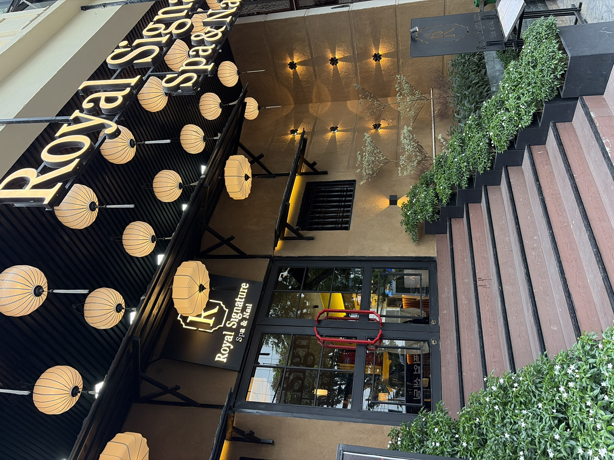 The image shows the entrance to "Royal Signature Spa & Nail" in Da Nang, Vietnam. A set of wooden stairs leads up to a glass door with red handles. The building's exterior is a warm brown color, and it is decorated with traditional Asian lanterns hanging from the ceiling. The entrance is flanked by lush green plants. Above the entrance, a prominent sign reads "Royal Signature Spa & Nail" in illuminated golden letters. Several wall-mounted lights cast a warm glow on the building's facade.
