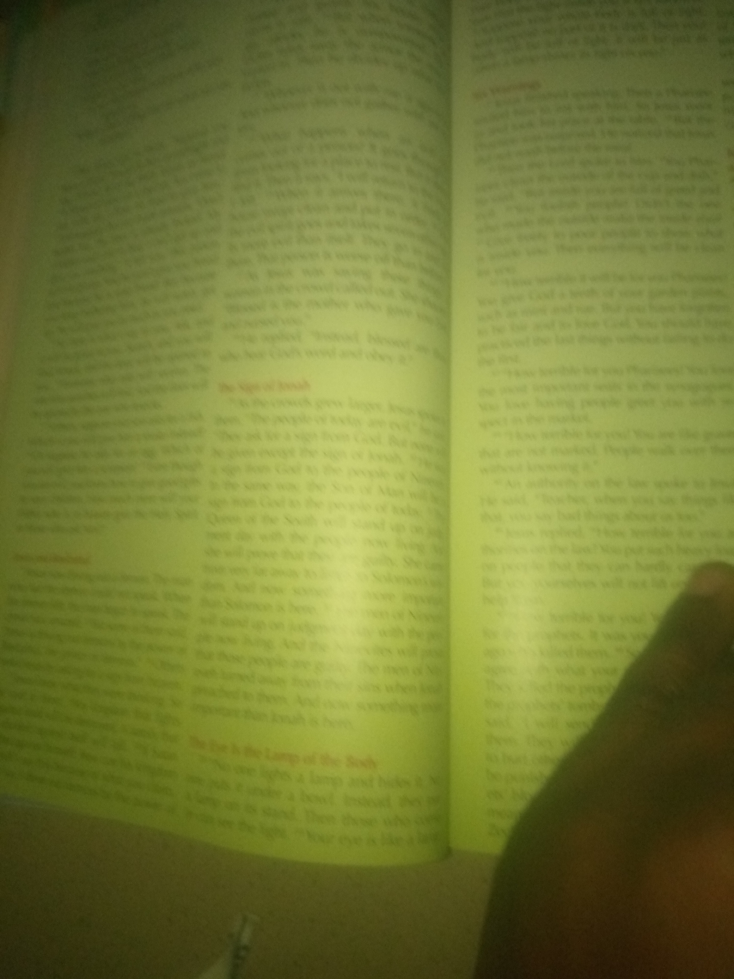 The image shows an open book with text on both pages. The book is yellowed, suggesting it is old. The text on the left page appears to be from a religious or historical text, possibly about a person named Jonah. The right page also contains text in a similar style. A dark, out-of-focus hand is visible in the lower right corner of the image, partially obscuring some of the text. The overall lighting is dim and yellowish. The scene is not identifiable as being in Jauro Yinu, Nigeria, or any specific city.