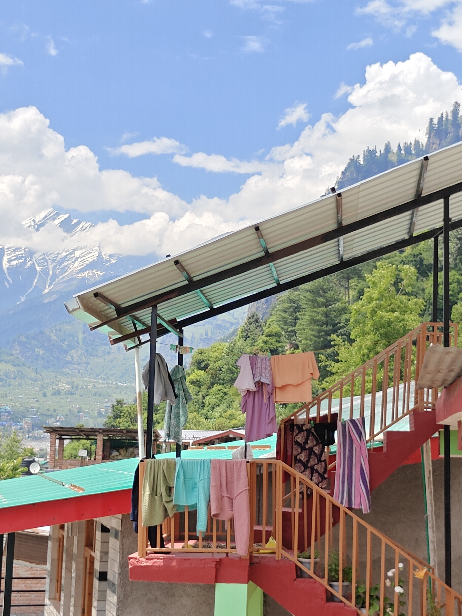 This image depicts an outdoor scene in Vashisht, India, on a clear day with scattered white clouds. The foreground features a red-painted staircase with wooden railings, on which various items of clothing are hung to dry. These include what appear to be sweaters, t-shirts, and pants in a variety of colors such as green, blue, pink, orange, purple, and striped patterns. Below the staircase, glimpses of buildings with green and red roofs are visible.
Behind the staircase and to the left, a covered structure with a corrugated metal roof supported by dark metal posts is visible. More clothing is hung from a line beneath this roof.
The background is dominated by lush green trees covering rolling hills, and further in the distance, majestic snow-capped mountains rise against the blue sky. The time of day appears to be daytime, likely late morning or afternoon, given the bright sunlight. The overall mood is serene and tranquil, conveying a sense of everyday life in a mountainous region. There is no visible text in the image.