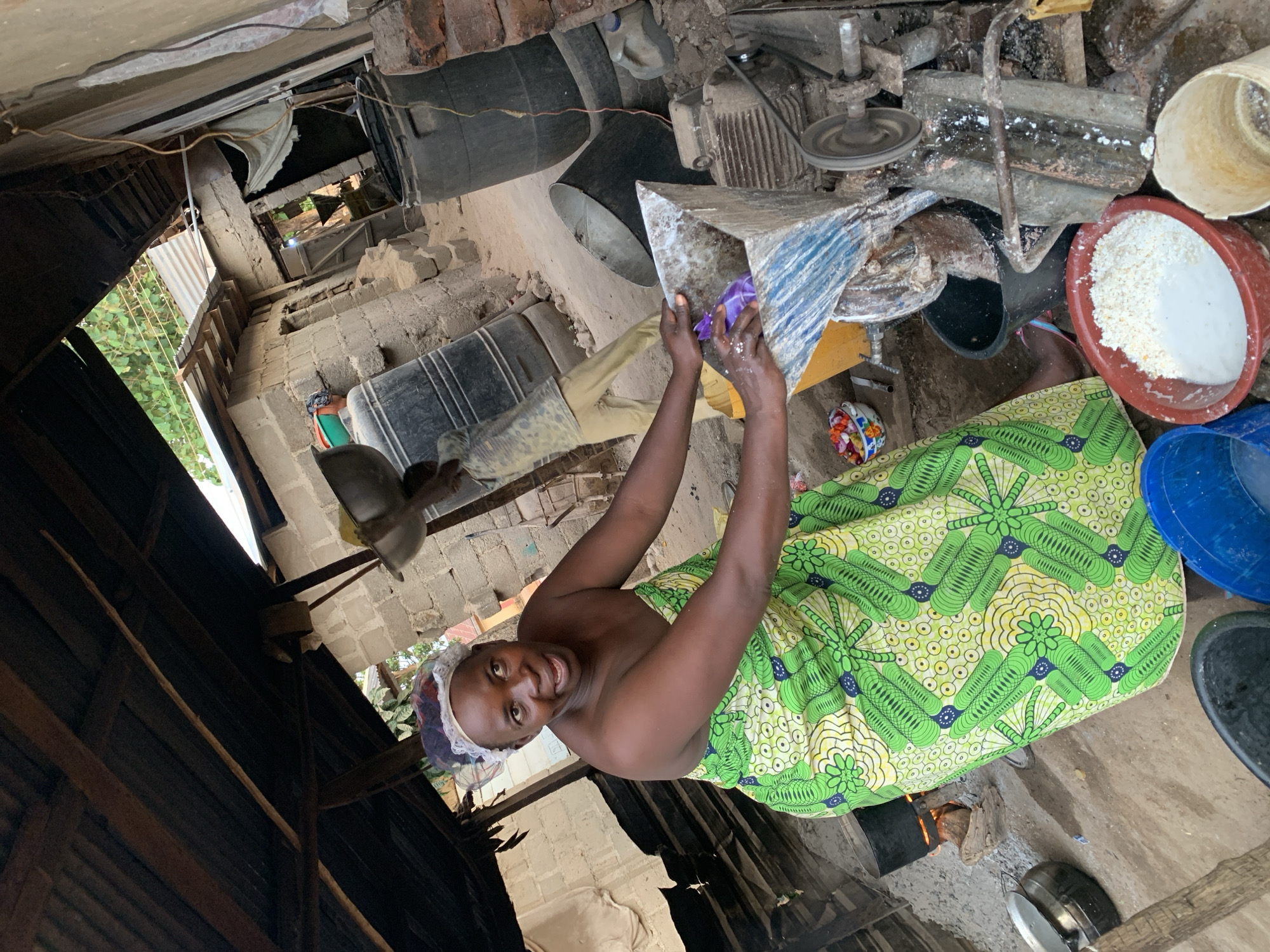 The image shows a woman processing cassava in a building in Agban, Nigeria. The woman, wearing a green patterned wrap and a hairnet, stands next to a machine with a metal funnel, and she is holding a purple cloth inside the funnel. To the right of the machine is a red bucket filled with a white, grainy substance. Around her are various containers, including black, blue, and white buckets. The structure she is in appears to be a rudimentary building with corrugated metal and brick walls and a partially open roof. An oven is on the ground next to her.