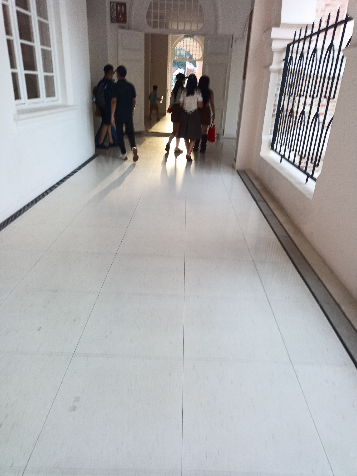 The image captures a wide-angle, low-perspective view of a brightly lit, tiled corridor in what appears to be a public building or institution. Sunlight streams in from the right, casting long shadows across the polished floor and illuminating the figures walking away from the camera.
In the foreground, the smooth, light-colored tiles dominate the scene, marked by dark grout lines that create a grid pattern. The corridor stretches into the distance, with a white wall on the left and a decorative black metal grate on the right.
Several people are visible further down the corridor, moving away from the viewer. On the left, two individuals in dark clothing walk side-by-side, with one carrying a backpack. To their right, a group of three individuals, appearing to be young women in uniforms, are walking together. One of them is carrying a red bag. A younger child in a light blue shirt is visible in the background, near an open doorway.
The architecture suggests a formal setting, possibly a school or government building, with arched doorways and tall windows. The overall impression is one of movement and light within an architectural space. The context of Kuala Lumpur, Malaysia, suggests this might be a historical or public building within the city.
