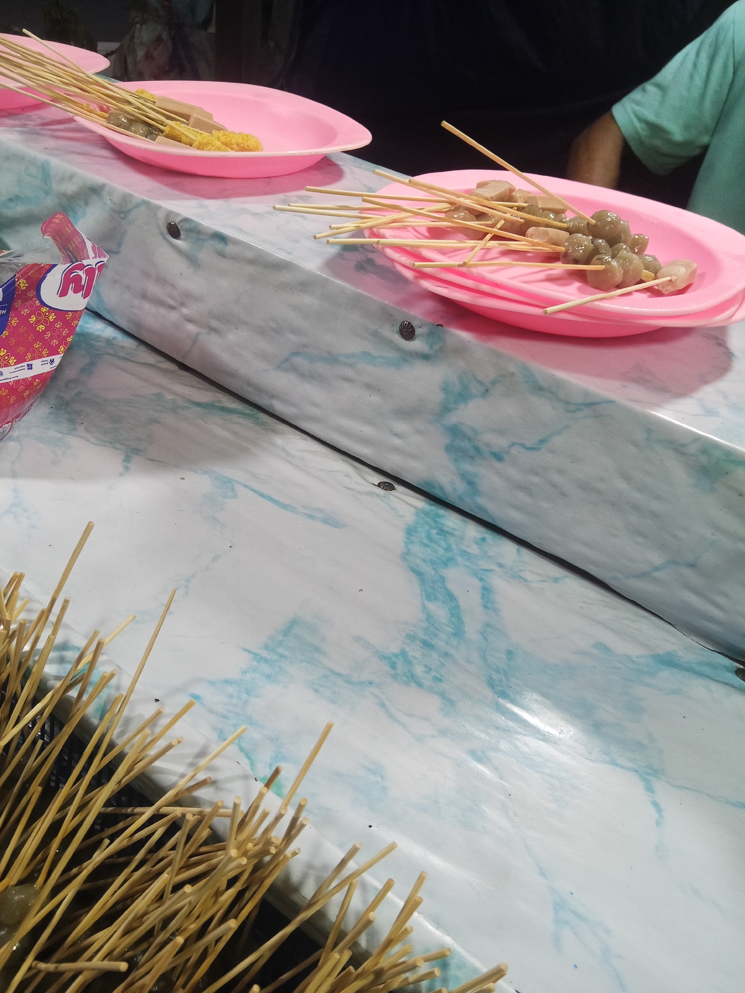 The image depicts a close-up view of food skewers on pink plastic plates, likely at a street food stall in Lubuklinggau, Indonesia. The skewers contain various items, including what appear to be meatballs, sausage, and corn. One plate is in focus, showing several skewers with meatballs and sausage slices. Another plate, partially visible, has skewers with corn and other unidentifiable food items. In the foreground, a bundle of unused wooden skewers is visible. The table surface has a marbled pattern with blue and white swirls. A person's arm in a green t-shirt is visible in the background, suggesting a vendor or customer. The overall setting appears to be an outdoor or semi-outdoor food stall, common in many Indonesian cities.