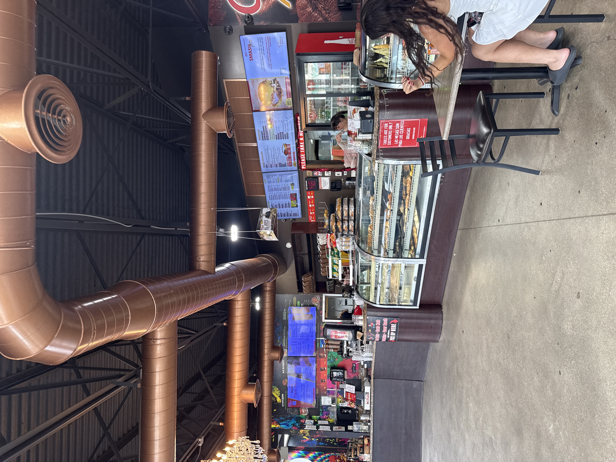 Here is a description of the image:
The image shows the interior of a cafe or restaurant. A person is seated at a high table near a counter where food is displayed in glass cases. Behind the counter, a worker is visible, likely serving customers. Menu boards are displayed above the counter. The cafe has a modern, industrial aesthetic with exposed pipes and ductwork on the ceiling.
Based on the menu boards for "Smashita" and what appears to be a sign stating "Tables are for customers only...Gracias, administracion," in conjunction with the food offerings displayed, it is possible this is a cafe or bakery restaurant serving Latin-inspired food in Miami Beach, Florida.