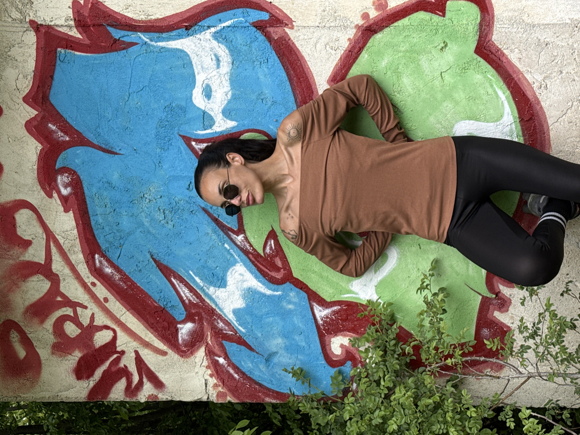 A young woman in sunglasses and wearing a brown long-sleeved shirt and black leggings lies on a graffiti-covered concrete structure. The graffiti consists of blue, green, red, and white paint. The wall is cracked. Vegetation grows at the base of the structure. The woman's tattoos are visible on her shoulder and chest. The location is Pleven, Bulgaria.
