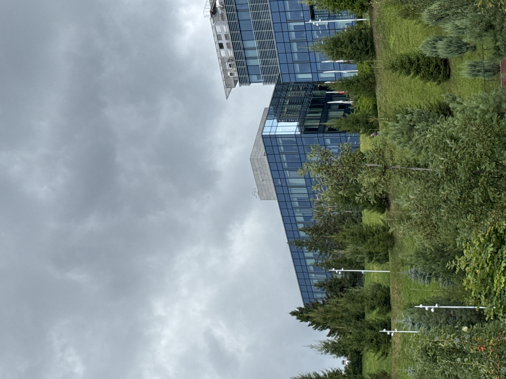 The image shows a modern office building with a glass facade, set against a cloudy sky. The building appears to be located on a green, hilly terrain, with various trees and shrubs in the foreground. There are multiple white lamp posts with multiple lights visible on the hillside. The architectural style of the building suggests a contemporary design, with clean lines and a prominent use of glass and metal. The surrounding vegetation indicates an outdoor, possibly park-like setting. The overall impression is one of a well-maintained corporate or commercial area. The image is likely captured in Kazan, Russia, given the context of the request, and the building could be a part of a business park or a corporate headquarters.
