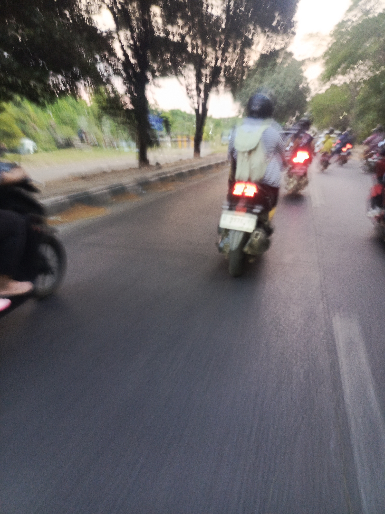 The image depicts a street scene in Sawotratap, Indonesia, characterized by motion blur. Several motorcycles are visible, traveling along a paved road lined with trees and greenery. In the foreground, a motorcycle rider is seen from behind, wearing a backpack and a patterned shirt. Their motorcycle has illuminated brake lights and a license plate. Other motorcycle riders are further ahead, also in motion. The environment suggests a daytime scene, possibly late afternoon given the lighting. The overall impression is one of active transportation on a road, with the blur indicating movement.