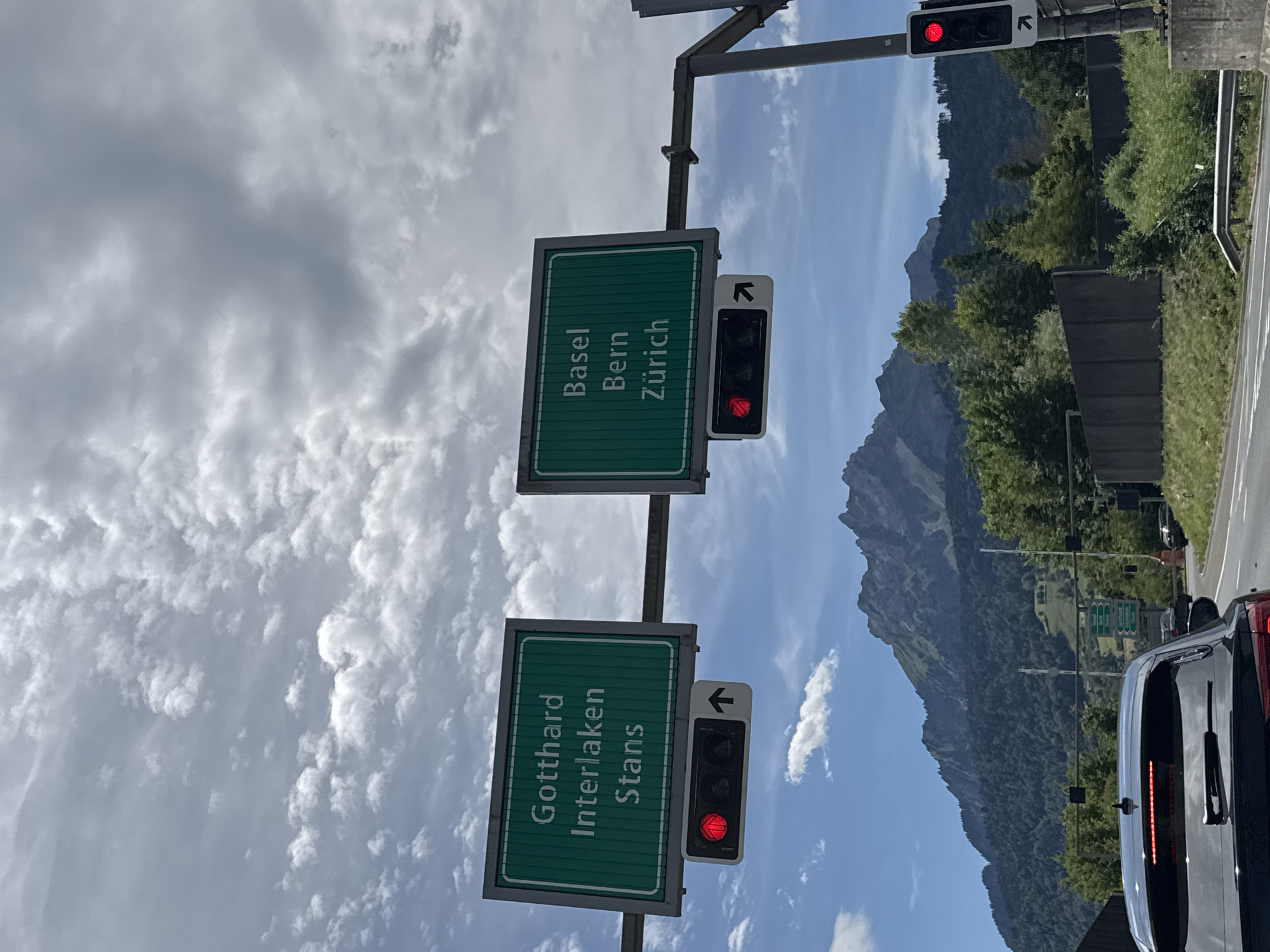 The image shows a street view in Kriens, Switzerland, featuring highway signage and traffic lights. Two green highway signs are visible. The left sign indicates directions to "Gotthard Interlaken Stans," and the right sign indicates directions to "Basel Bern Zürich." Below each sign, there is a traffic light with a red signal illuminated. An upward-pointing arrow is also visible on the right side of each traffic light housing. In the foreground, the rear of a black car is visible, and further in the background, a mountainous landscape with dense forest cover can be seen. The sky is partly cloudy.