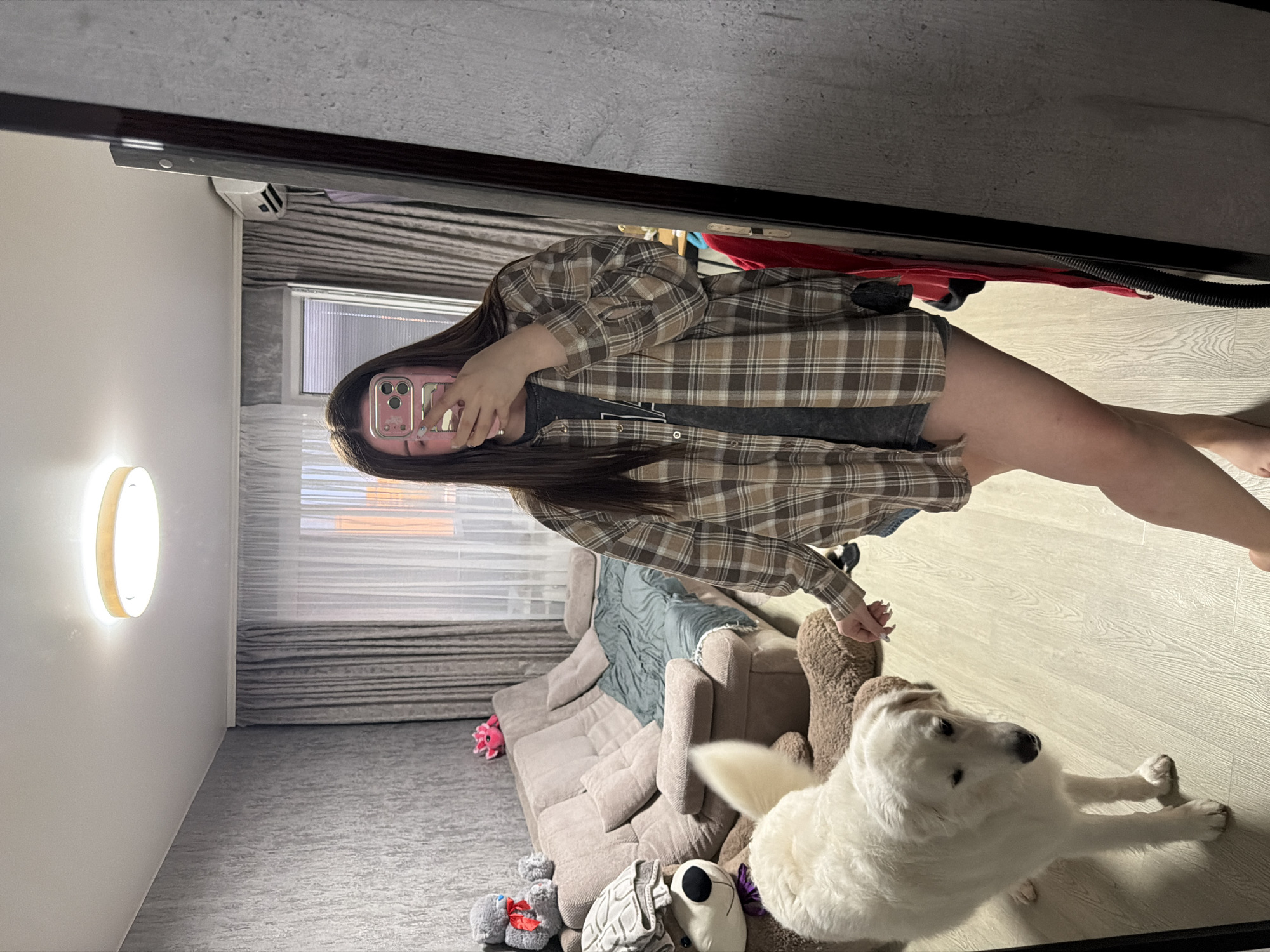 A medium shot captures a young woman standing in a domestic interior, reflected in a large mirror on the right side of the frame. The woman has long, dark hair and is holding a pink smartphone to take the photo. She is wearing a loose-fitting, brown and beige plaid shirt over a dark t-shirt and grey shorts. Her left hand is extended towards a large, fluffy white dog that is standing on a light-colored wooden floor in front of her. The dog is looking down. Behind the dog and to the left, a large beige sofa is visible with several pillows and a blue blanket draped over it. Various stuffed animals, including a large teddy bear, are placed on and around the sofa. The background shows a grey textured wall, a window with grey curtains and sheer white drapes, and a ceiling light fixture. An air conditioning unit is mounted high on the wall near the window. The overall environment appears to be a living room.