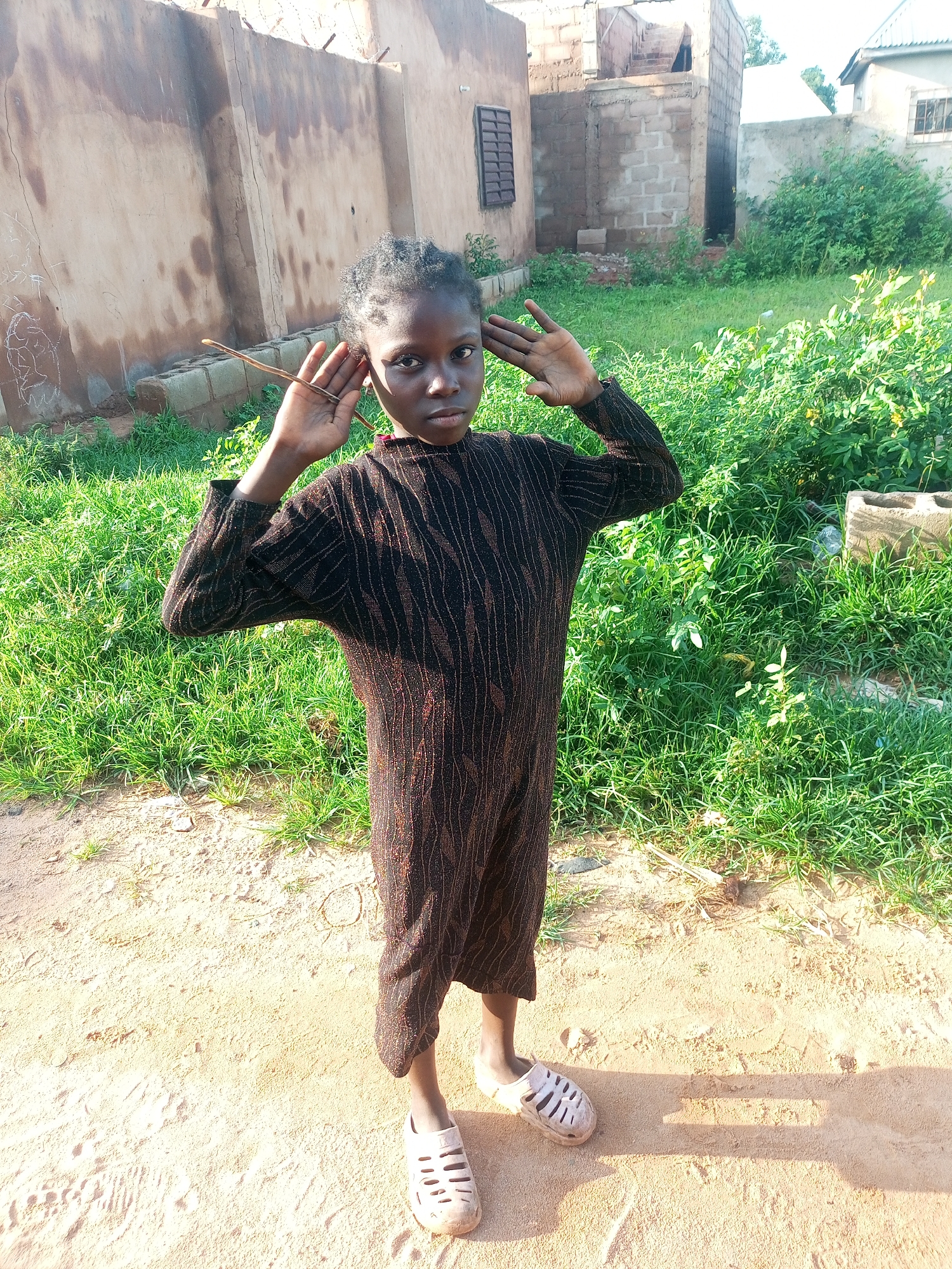 A young girl stands in a sandy outdoor area with a building and greenery in the background, possibly in Tempure, Nigeria. She is wearing a long-sleeved, dark-colored dress with a patterned design. Her arms are raised, with her hands near her ears, and she is looking directly at the camera. Her hair is dark and styled in short braids. She is wearing light-colored, open-toed sandals. The ground is a mix of sand and dirt, with sparse green grass and some bushes in the background. A partially constructed building with brickwork and a barred window is visible behind her. The lighting suggests it is daytime.