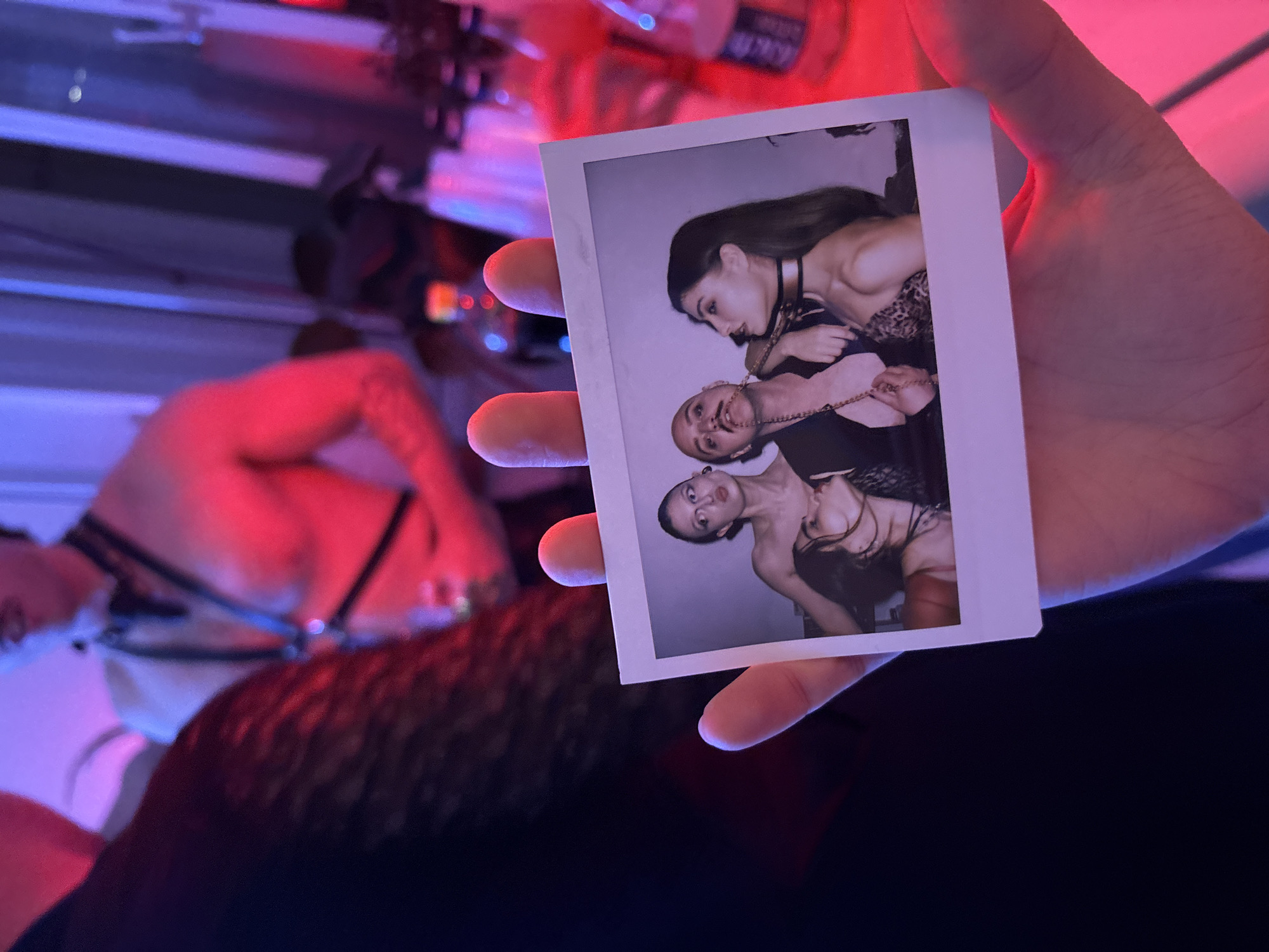 A person's hand holds a Polaroid photo featuring three individuals, all women. The woman at the top of the Polaroid wears a black choker and a leopard-print top, gazing down at the other two. The middle woman has light hair and is connected to the first by a gold chain, smiling toward the camera. The woman at the bottom of the Polaroid also appears to be connected by the same chain.
In the background, slightly blurred, a person is seen wearing leather restraints or a harness-like garment. The overall lighting is low, with a pink and blue color cast. The location cannot be determined to be in Moscow, Russia.