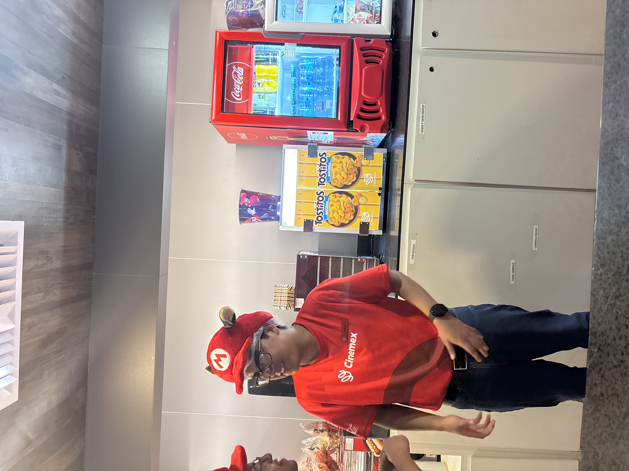 The photograph shows two people wearing "Mario Bros." hats and red "Cinemex" employee shirts, standing in front of a snack bar, likely in a Cinemex movie theatre. The snack bar features a Coca-Cola branded refrigerator, and boxes of "Tostitos Nachos." The location may be in Puebla, Mexico due to the Cinemex branding.
