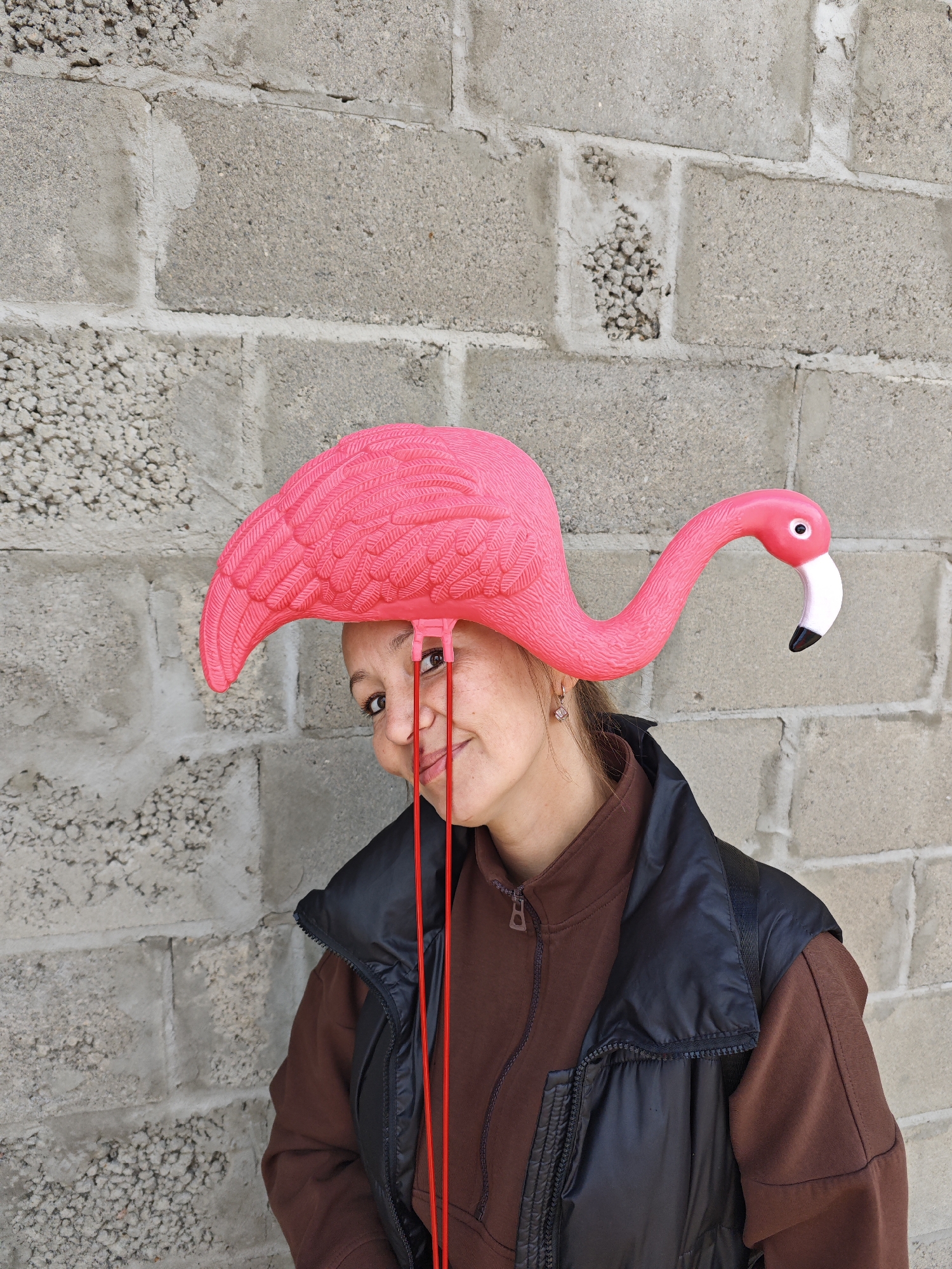 A middle-aged woman stands in front of a cinder block wall, wearing a brown jacket and a black vest. On her head, she has a bright pink plastic flamingo. The scene is located in the city of Chaikovsky, Russia. Two red strings hang down in front of her face.