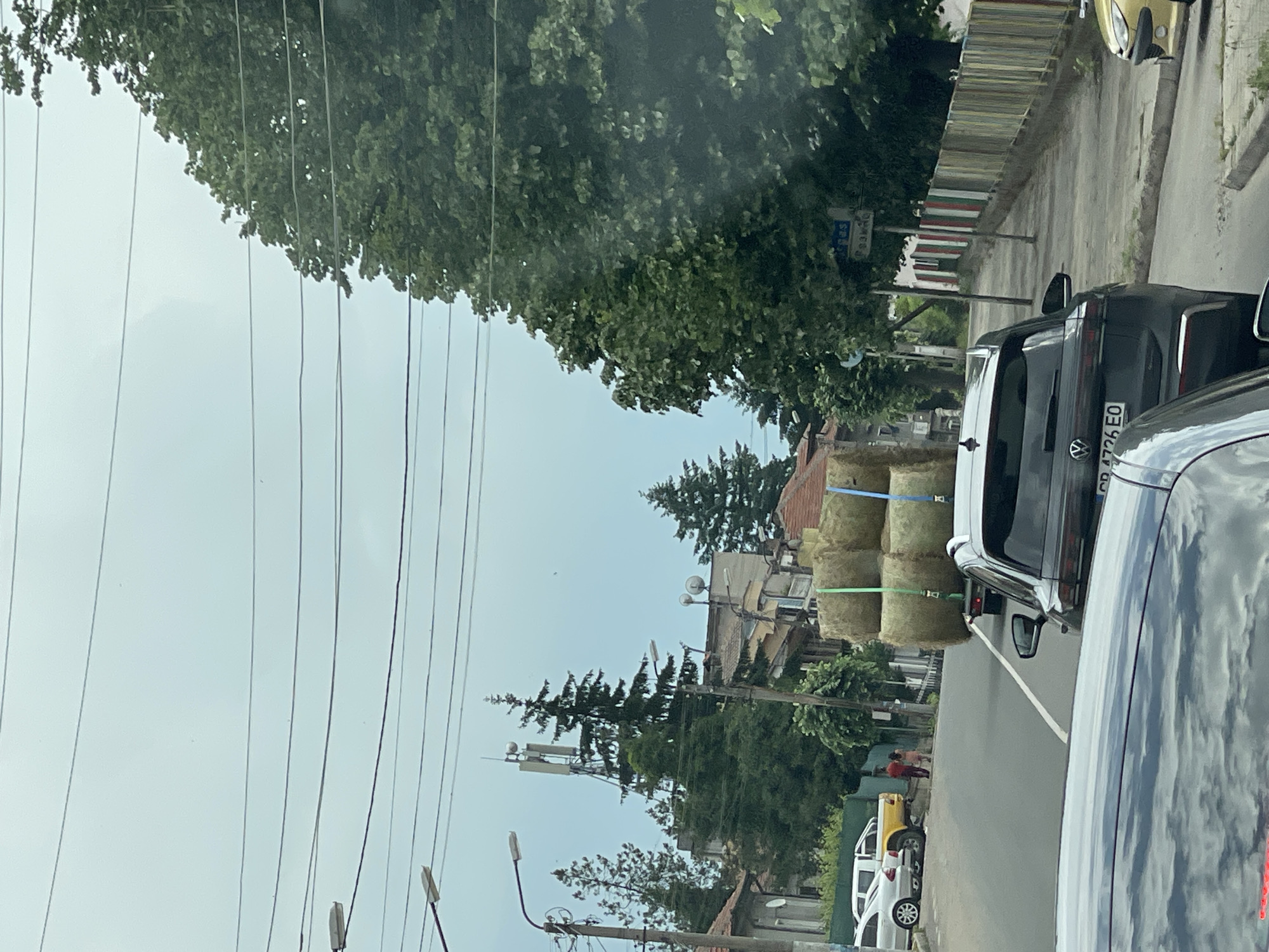The image depicts an overcast daytime street scene in Rozino, Bulgaria, observed from within a vehicle.
In the foreground, the partially visible hood and windshield reflections of a dark vehicle are present. Directly ahead, a dark grey Volkswagen SUV, bearing the Bulgarian license plate "PB 4726 EO" (indicating Plovdiv district), is in a traffic lane. Attached to the rear of the SUV is a flatbed trailer. The trailer is loaded with three large, round hay bales, secured with visible green and blue straps.
To the left of the road, a yellow pickup truck and a silver sedan are parked. Two individuals are visible on the sidewalk near the yellow pickup, one in a red shirt and the other in a lighter-colored top, standing or walking away from the camera. The road surface is asphalt, marked with a solid white line.
On the right side of the street, a sidewalk runs alongside a multicolored picket fence, featuring vertical segments painted green, white, and red. A blue street sign, mounted on a pole, is visible on this sidewalk, displaying text in Cyrillic; the top line reads "РАЗГРАД" (Razgrad) and the bottom line "УЛИЦА РОДИНА" (Rodina Street).
The background consists of various residential buildings, many with traditional tiled roofs, interspersed with dense green trees of both coniferous and deciduous types. A prominent cellular tower with multiple antennae stands tall in the distance, situated behind some evergreen trees and seemingly atop a building. Numerous overhead utility wires traverse the upper portion of the scene, strung between utility poles equipped with streetlights. The sky is a uniform light grey, indicating overcast conditions.