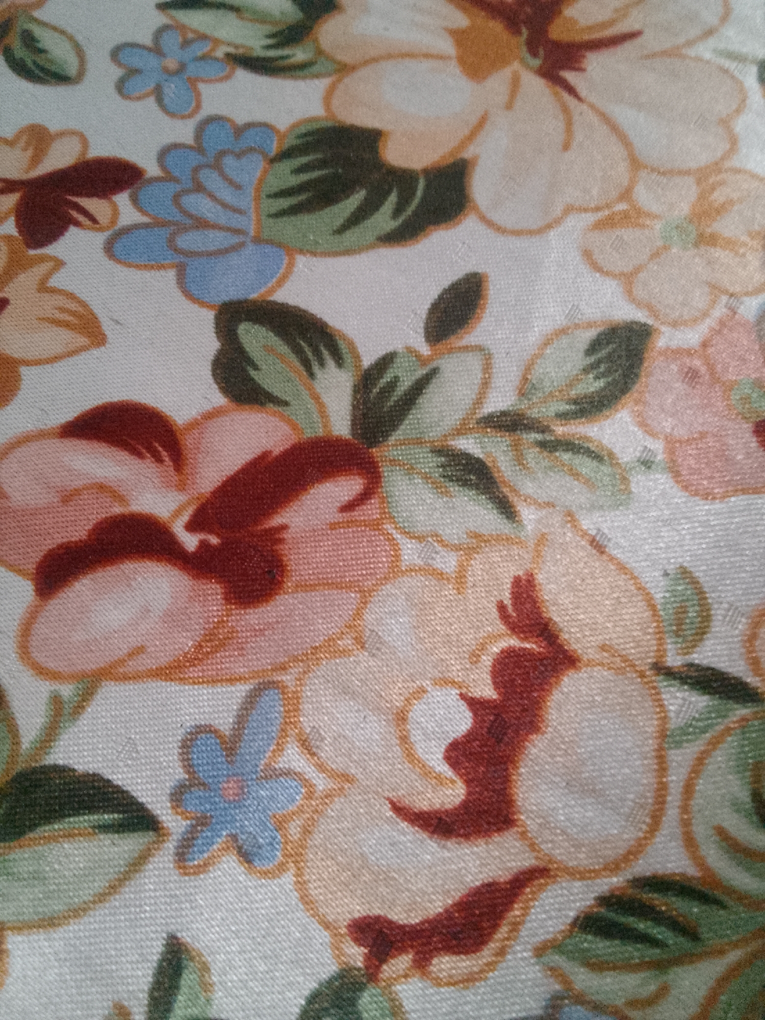 The image displays a close-up of a fabric with a floral pattern. The pattern features large, stylized flowers in shades of cream, peach, and pink, with accents of deep red in the centers and along the edges of some petals. Green leaves with dark outlines are interspersed throughout the design, adding depth and contrast. Smaller blue flowers with pink centers are also present. The fabric appears to have a subtle sheen, suggesting it might be satin or a similar material. The pattern is densely arranged, covering the entire visible surface of the fabric without any discernible background elements other than the fabric weave itself. There are no people, actions, or interactions depicted in the image. The image is a still photograph of a textile. This image is not located in the city of Karangan, Indonesia.