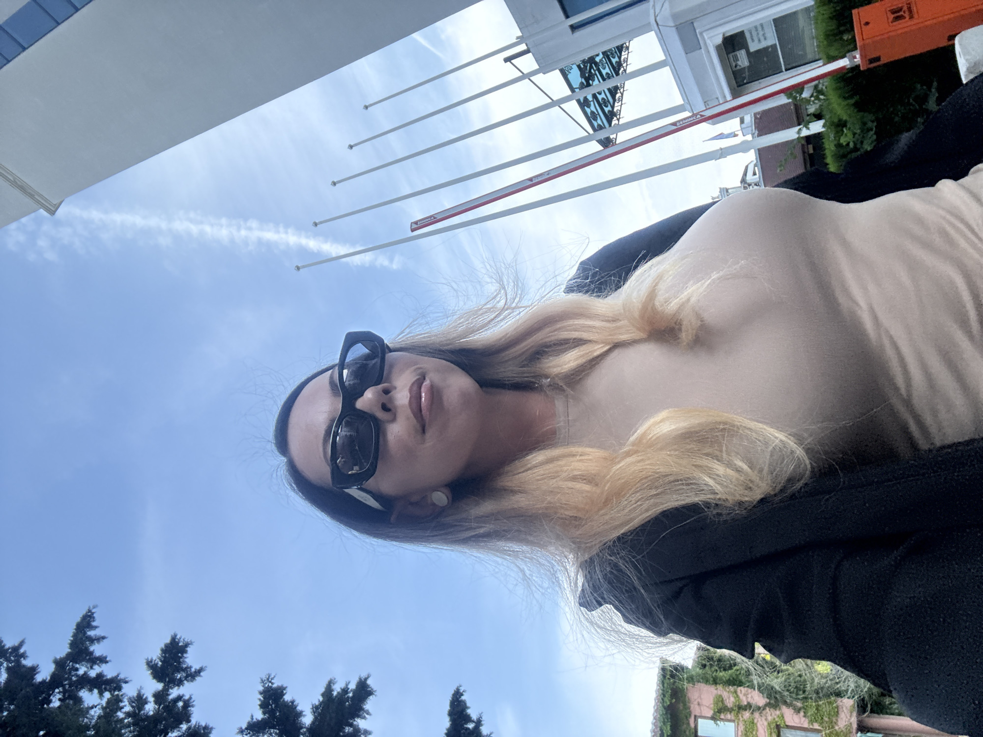 A low-angle, close-up selfie shows a woman wearing large, black, rectangular sunglasses and a light beige, form-fitting top under a dark jacket. She has long, blonde hair that is windswept. In the background, a bright blue sky with wispy clouds is visible. To the left, the dark green foliage of a tree hangs into the frame. To the right, a tall, white building with blue-tinted windows rises vertically. Several tall, thin white flagpoles stand in a row in front of the building, with a red and white barrier gate between them. Further in the background, a pink building covered in green ivy can be seen. This scene is likely in Nesebar, Bulgaria, given the architectural style and the presence of flagpoles often seen at official or tourist sites. The woman is looking upwards and slightly to the right, away from the camera.